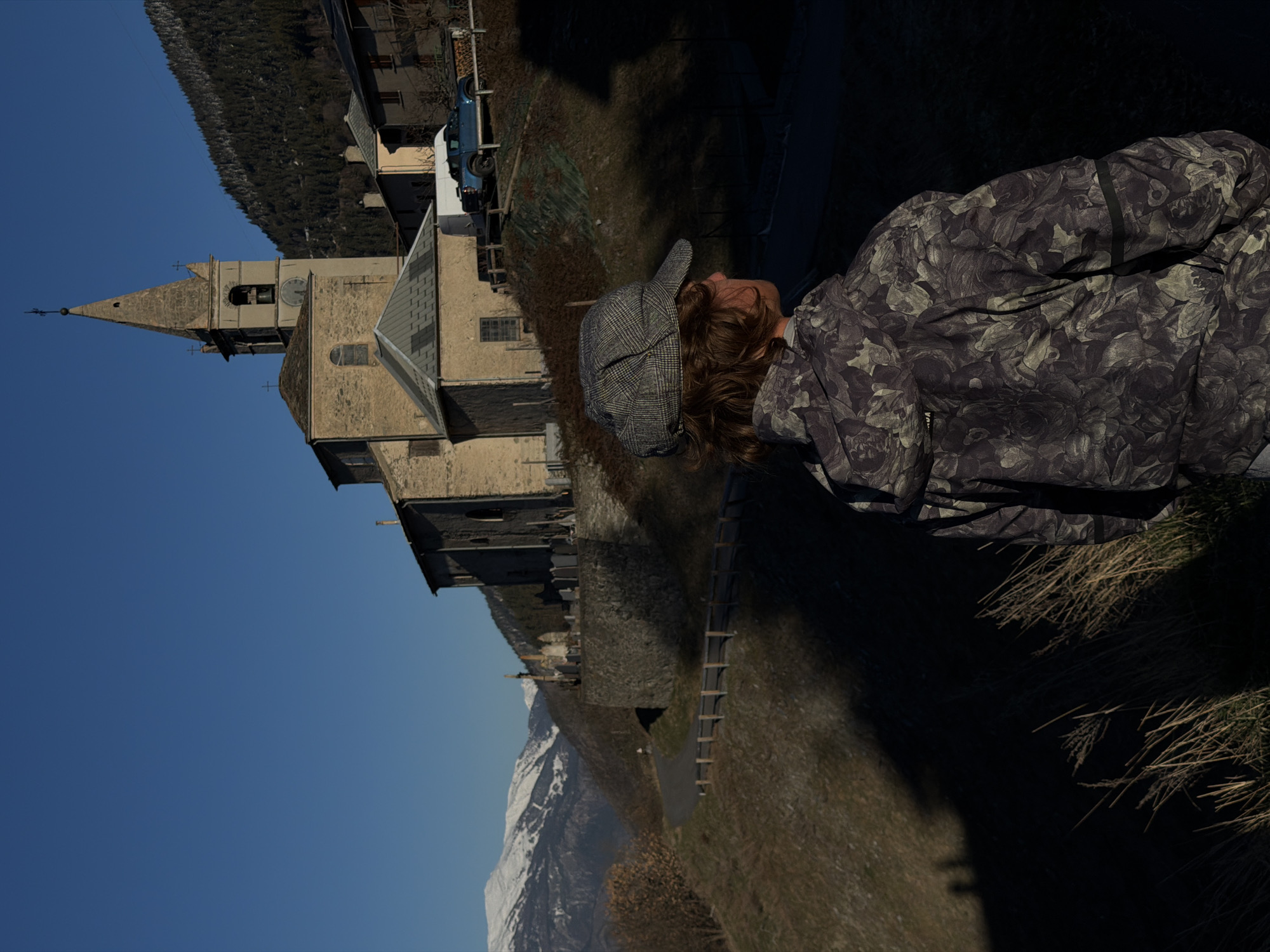 A person wearing a cap and a patterned jacket is looking at the Saint-André church, located in Saint-André, France. The church has a tall spire and a stone exterior. Mountains are visible in the background. A blue and white vehicle is parked near the church. A small road is adjacent to the church, and a grass-covered slope is in the foreground.