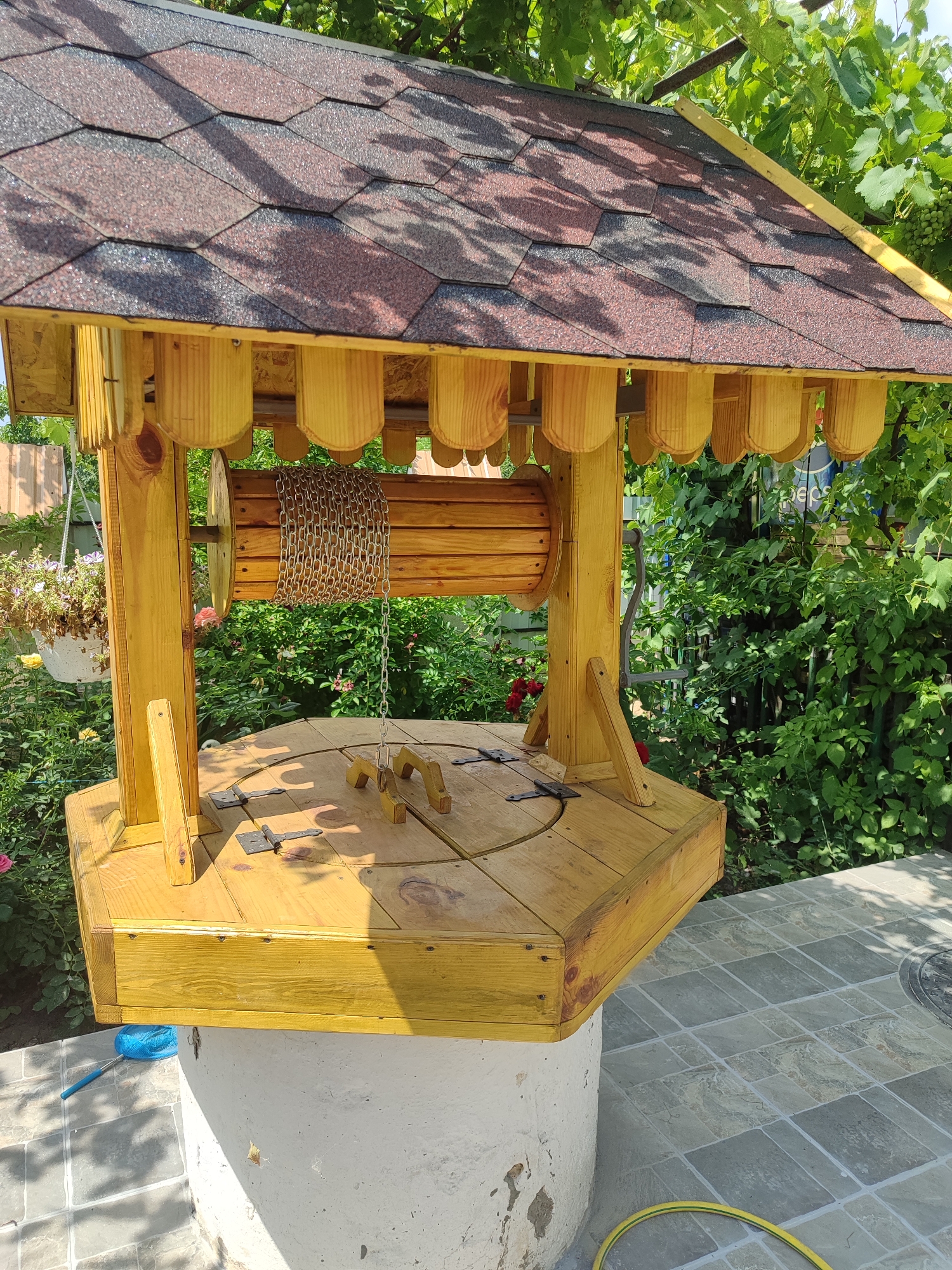 An octagonal wooden well structure with a shingled roof is situated outdoors in a garden. The well has a wooden drum with a metal chain coiled around it. The top of the well is covered with wooden planks. The structure is painted yellow and appears to be recently constructed. The background is filled with lush green foliage and flowering plants. Sunlight filters through the leaves, casting shadows on the well. The ground around the well is paved with stone tiles.