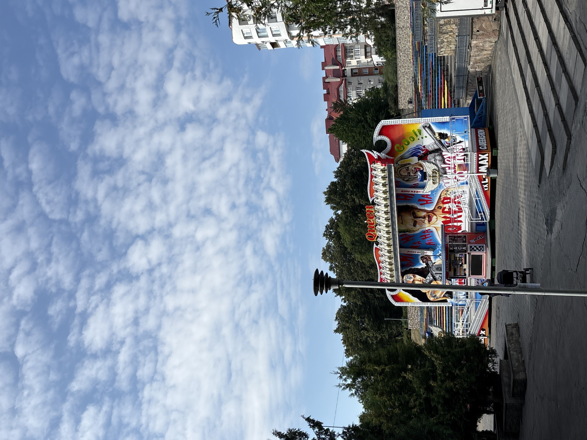 The image captures a bright, cloudy day in Ternopil, Ukraine, with the main subject being a carnival ride that appears to be themed around a "Queen" show. The ride features vibrant graphics, including images of characters resembling DC Comics' Harley Quinn and the Joker, along with a figure identified as Wonder Woman. The word "Queen" is prominently displayed in large, stylized lettering at the top of the ride.
In the foreground, a paved area with some stone steps leads towards the ride. A lamppost stands to the left of the ride. A person pushing a stroller is visible in the mid-ground, walking away from the ride. Behind the ride, a densely wooded area with tall trees forms a backdrop, and further in the distance, several residential buildings with red-tiled roofs and one taller white building are visible against the blue sky dotted with fluffy white clouds. The overall scene suggests an amusement or entertainment area within the city.