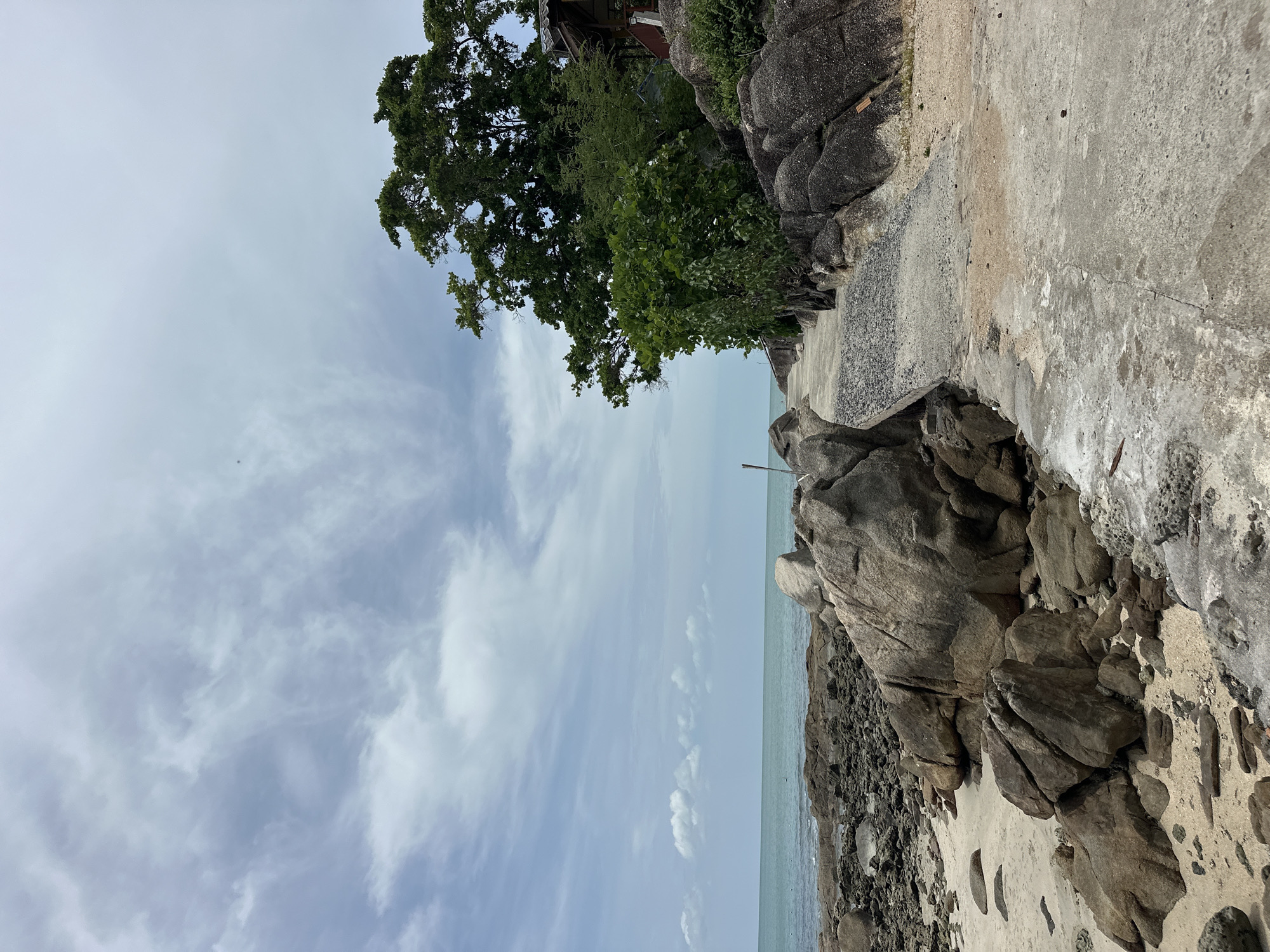 This is a ground-level, eye-level, medium shot capturing a weathered concrete pathway leading along a rocky coastline. The pathway is cracked and stained, with areas of exposed gravel. To the left, the pathway runs alongside a beach dotted with large, smooth, grey and brown rocks. Beyond the rocks, the turquoise ocean stretches to the horizon under a cloudy sky. To the right of the pathway, a steep incline is covered in large boulders and lush green foliage, including a tall, mature tree. A small, rustic wooden structure with a dark roof is partially visible amongst the trees. The scene appears to be a secluded coastal area, likely in a tropical region given the vegetation. The location is the beach of the northern part of Koh Samui island, Thailand, in the Ko Samui District.