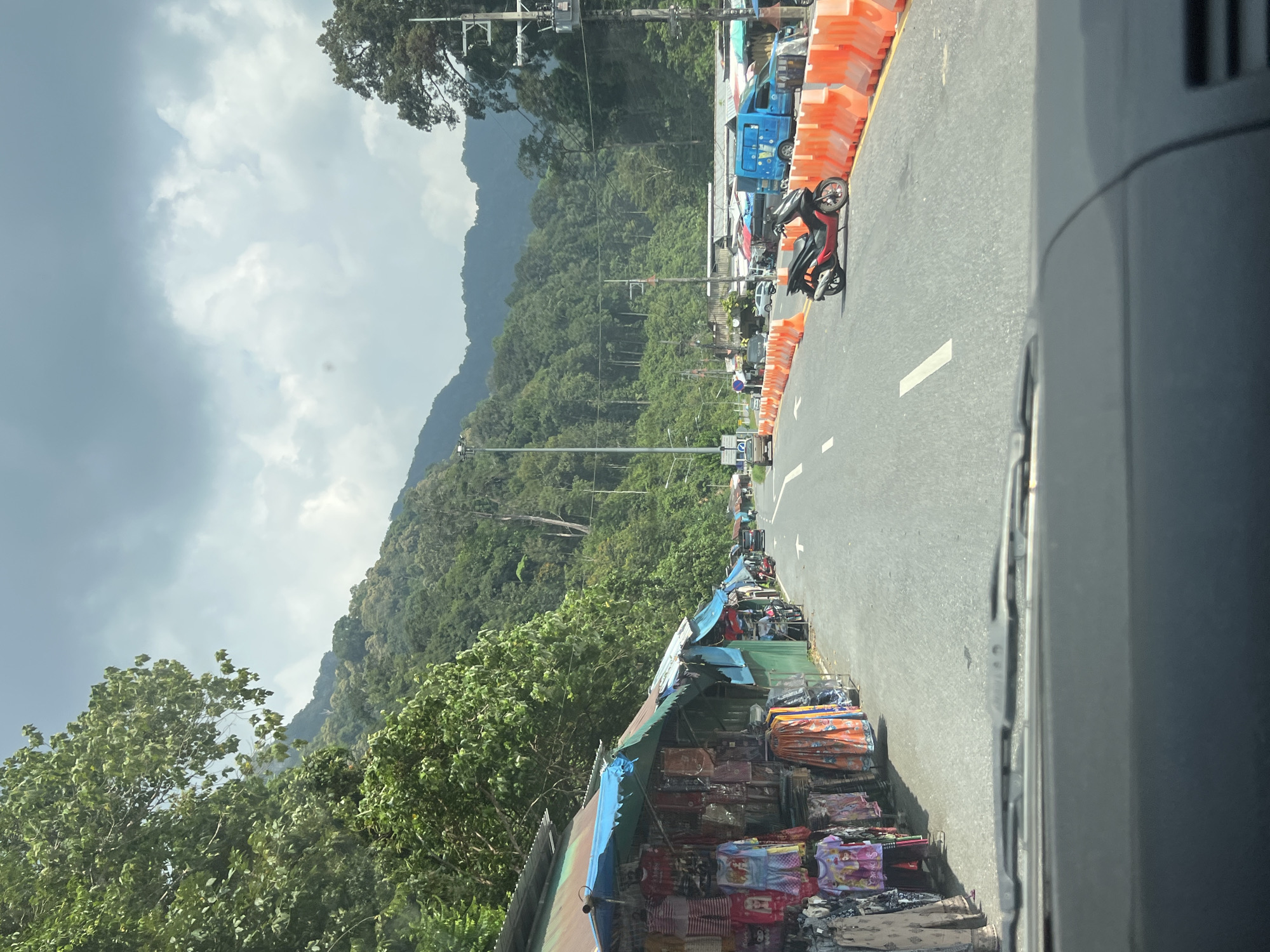 The image depicts a roadside scene in Tambon Wang Prachan, Thailand. The foreground shows the interior of a vehicle, with the windshield wipers visible. The scene outside consists of a two-lane road with a motorcycle parked on the right side, separated by orange traffic barriers. On the left side of the road are several market stalls selling clothing. In the background, a lush, green mountainside is covered in dense forest. The sky is overcast with dramatic clouds. The overall atmosphere suggests a commercial area situated along a scenic, mountainous route.