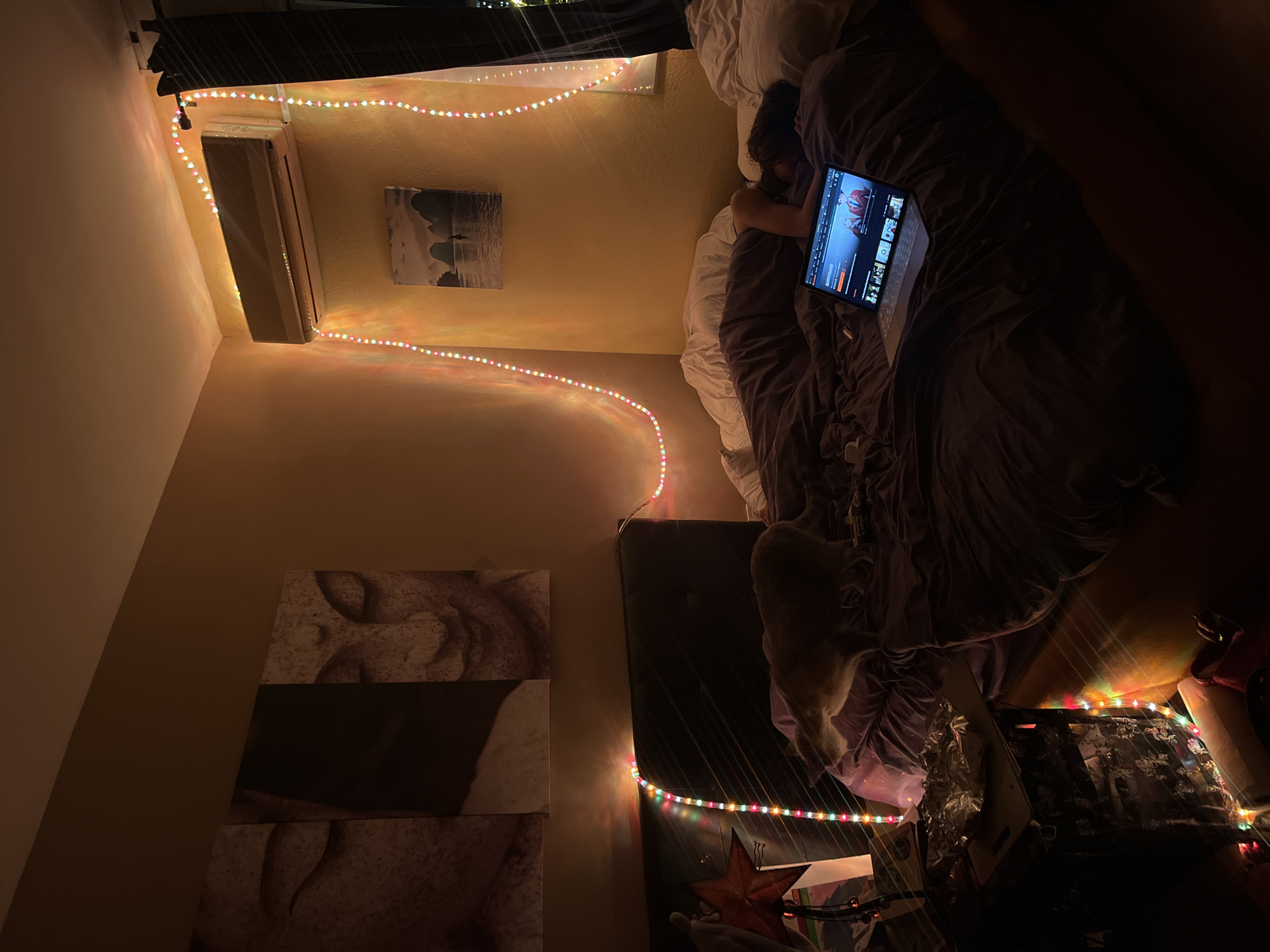 The image, taken from an elevated perspective and rotated, presents a cozy, dimly lit bedroom scene at night. A person is lying on their side under dark purple bedding on a large bed, attentively watching a laptop. The laptop screen displays what appears to be a streaming service interface, showing multiple show or movie thumbnails, with a prominent one featuring red animated or cartoon characters. A white cat is also visible on the bed, positioned towards the foot of the bed.
Colorful LED string lights, showcasing red, green, blue, and yellow dots, are strung along the room's walls, providing the primary illumination and casting soft glows on the light brown/tan walls. The lights run along the wall where an air conditioning unit is mounted, and then down a side wall.
Above the bed, a rectangular piece of wall art depicts a dark landscape scene. Further down the side wall, three stacked rectangular art pieces display abstract, textured patterns. Below the string lights on the side, a dark nightstand holds a decorative red star and other various objects. Through a window visible at the top, distant city lights confirm the nighttime setting in Barcelona, Spain. The overall ambiance is one of comfortable relaxation.