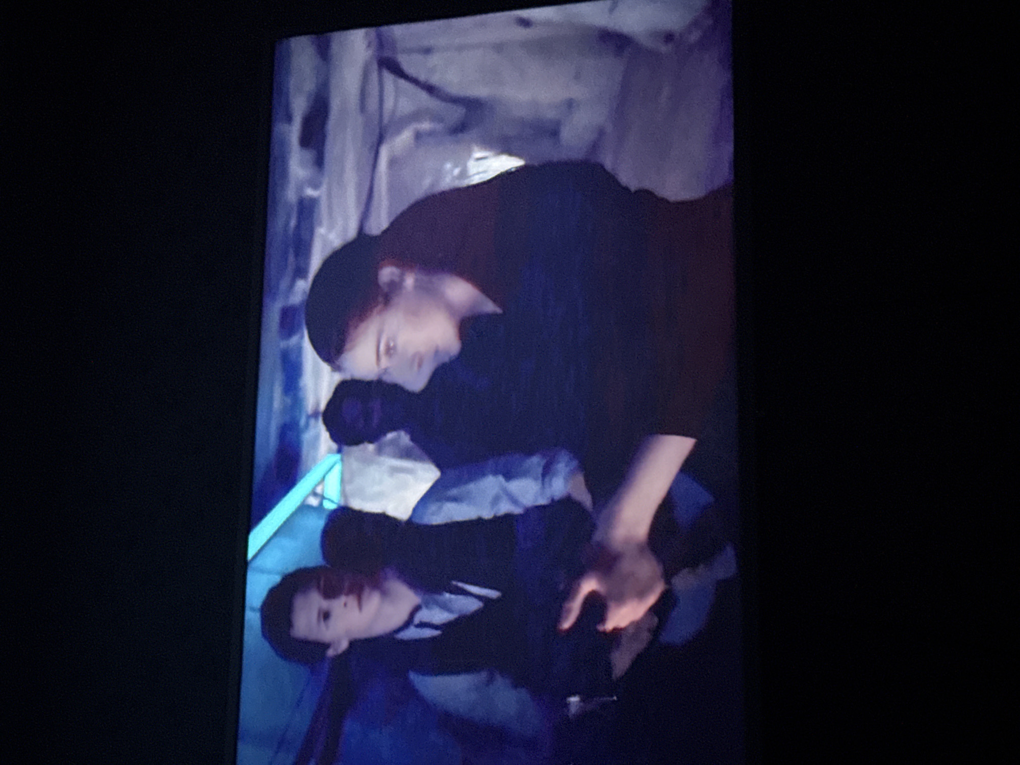 The image shows two individuals indoors. One is lying down with another person leaning over them. Both appear to be in a dimly lit, enclosed space, possibly a tent or shelter. The environment seems to be rudimentary, potentially a temporary or makeshift setting. There is a pale, possibly reflective, surface behind the people, but the details are obscured. The action suggests assistance or caregiving. This information does not include specifics tying it to Mytishchi, Russia.