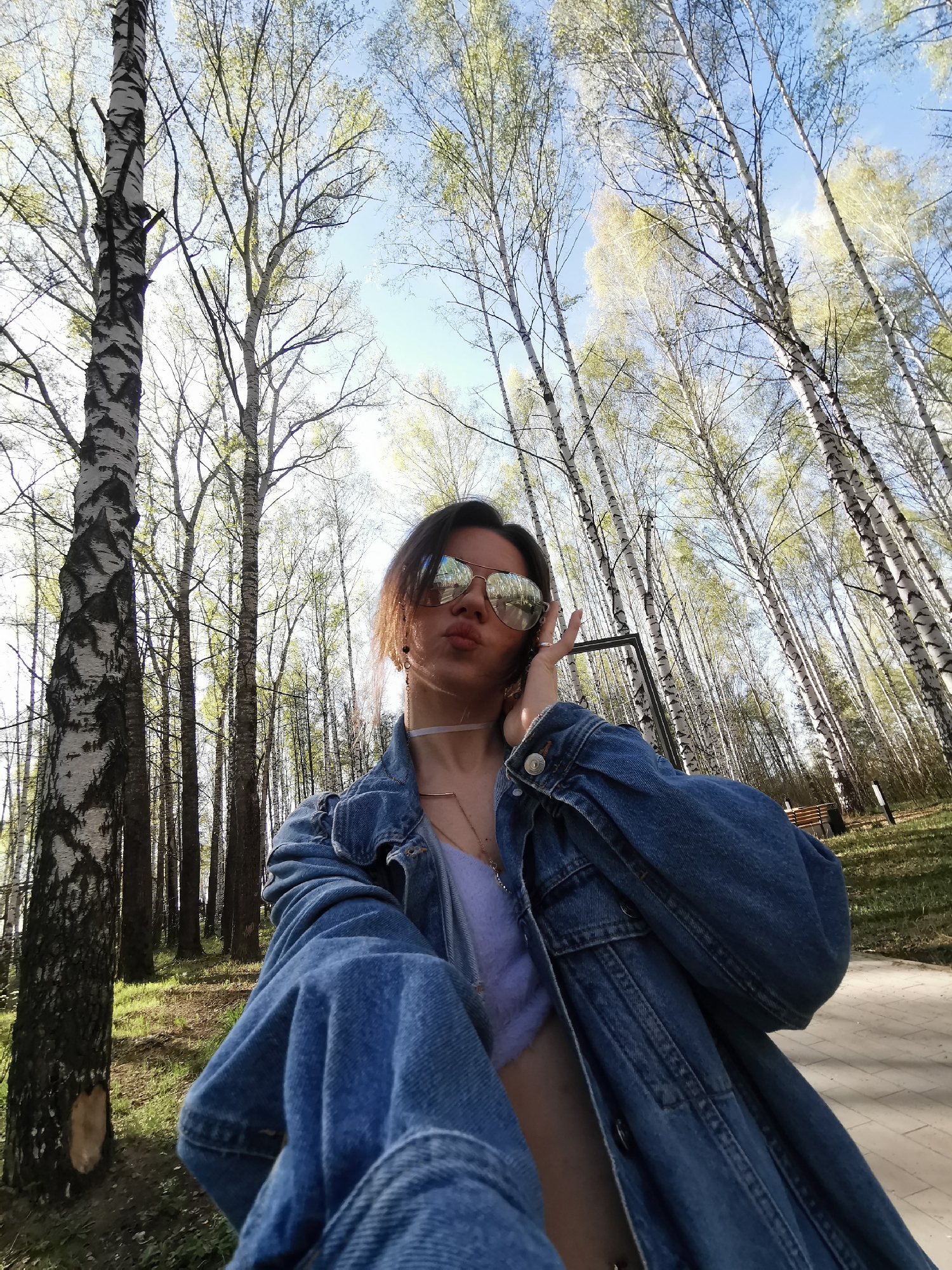 A low-angle shot captures a woman outdoors. The setting appears to be a park, evidenced by numerous birch trees with their distinctive white bark visible in the background. A paved path is partially seen on the right.
The woman is wearing a blue denim jacket, a light-colored top, and sunglasses. Her hair is dark and styled casually. She is holding up her hand in a gesture near her face. Her expression is a pout.
The sky is visible through the trees. A bench is partially visible.