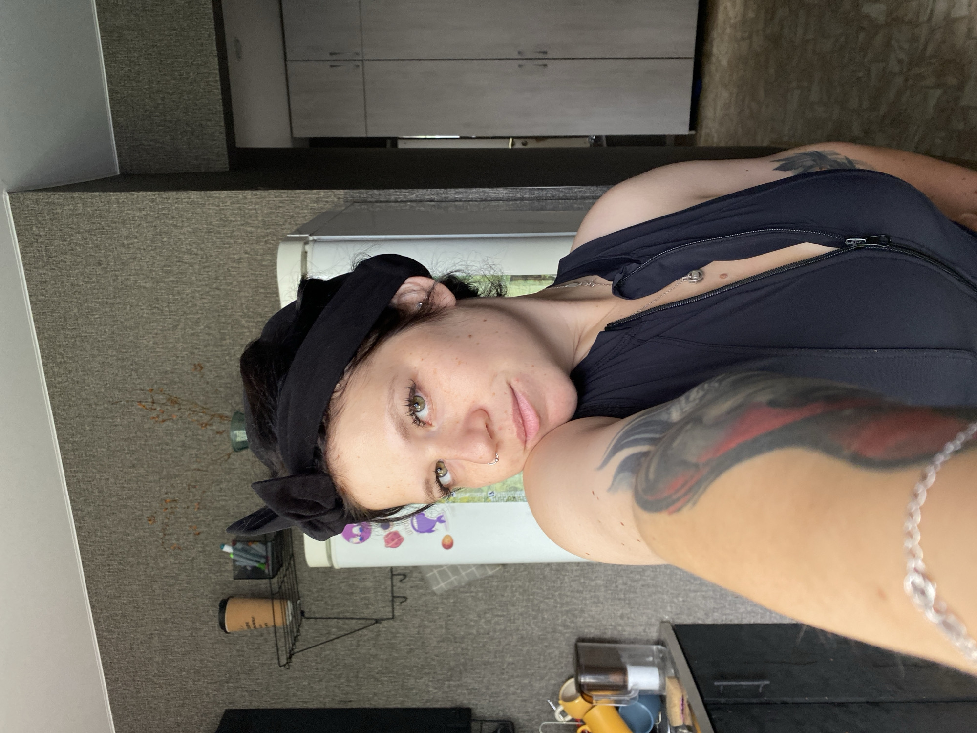 A young woman is taking a selfie in what appears to be a kitchen or living area. She has dark hair pulled back with a black bandana and is wearing a black sleeveless top with a zipper. She has a nose ring and a tattoo on her right arm. In the background, there is a refrigerator with stickers, a shelf with cups, and cabinets. The overall lighting suggests it is daytime. The scene is indoors, likely in a residential setting in Chaikovsky, Russia.