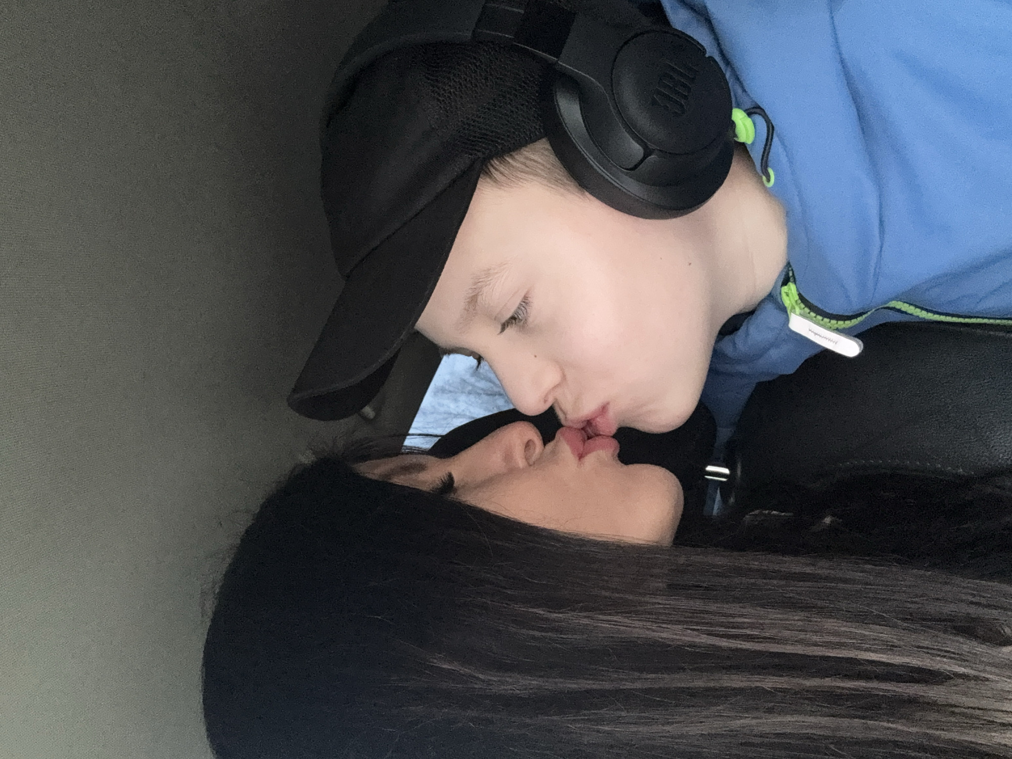 A boy wearing a black baseball cap and JBL headphones and a woman with long dark hair are seen kissing. The setting appears to be inside a car. The specific location of Chaikovsky, Russia is not discernible from the image.