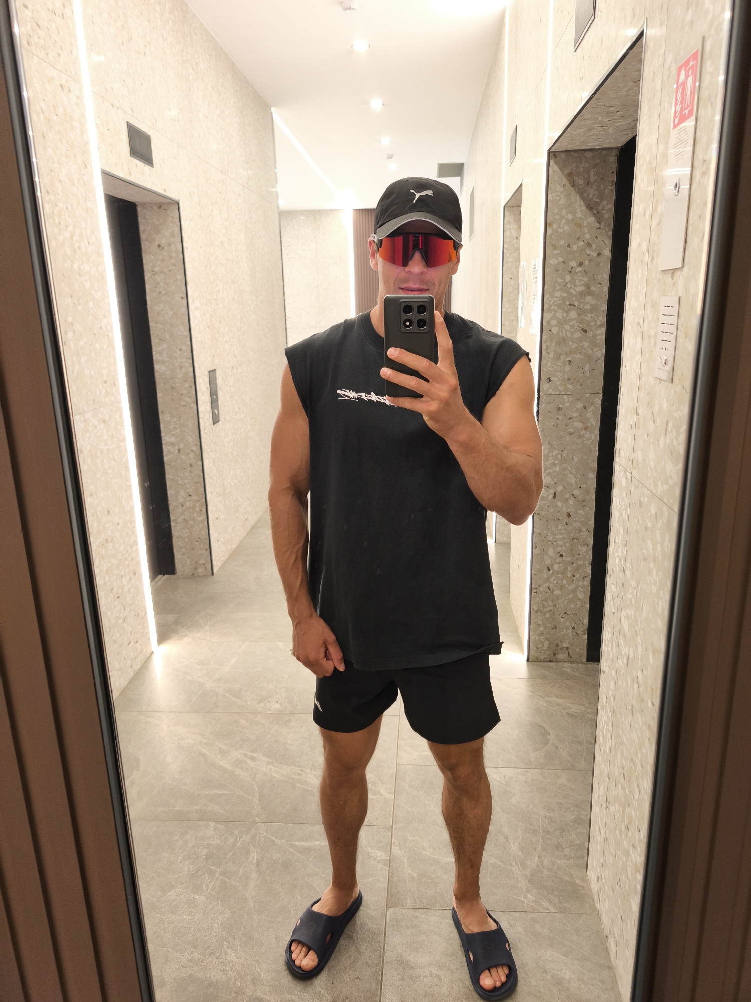 A man is taking a selfie in a hallway with two elevators. He is wearing a black sleeveless shirt, black shorts, a black baseball cap, red-tinted sunglasses, and blue slides. He is holding a smartphone in his right hand. The hallway has beige tiled walls with vertical strip lighting and grey marble-patterned flooring. The elevators are visible in the background.