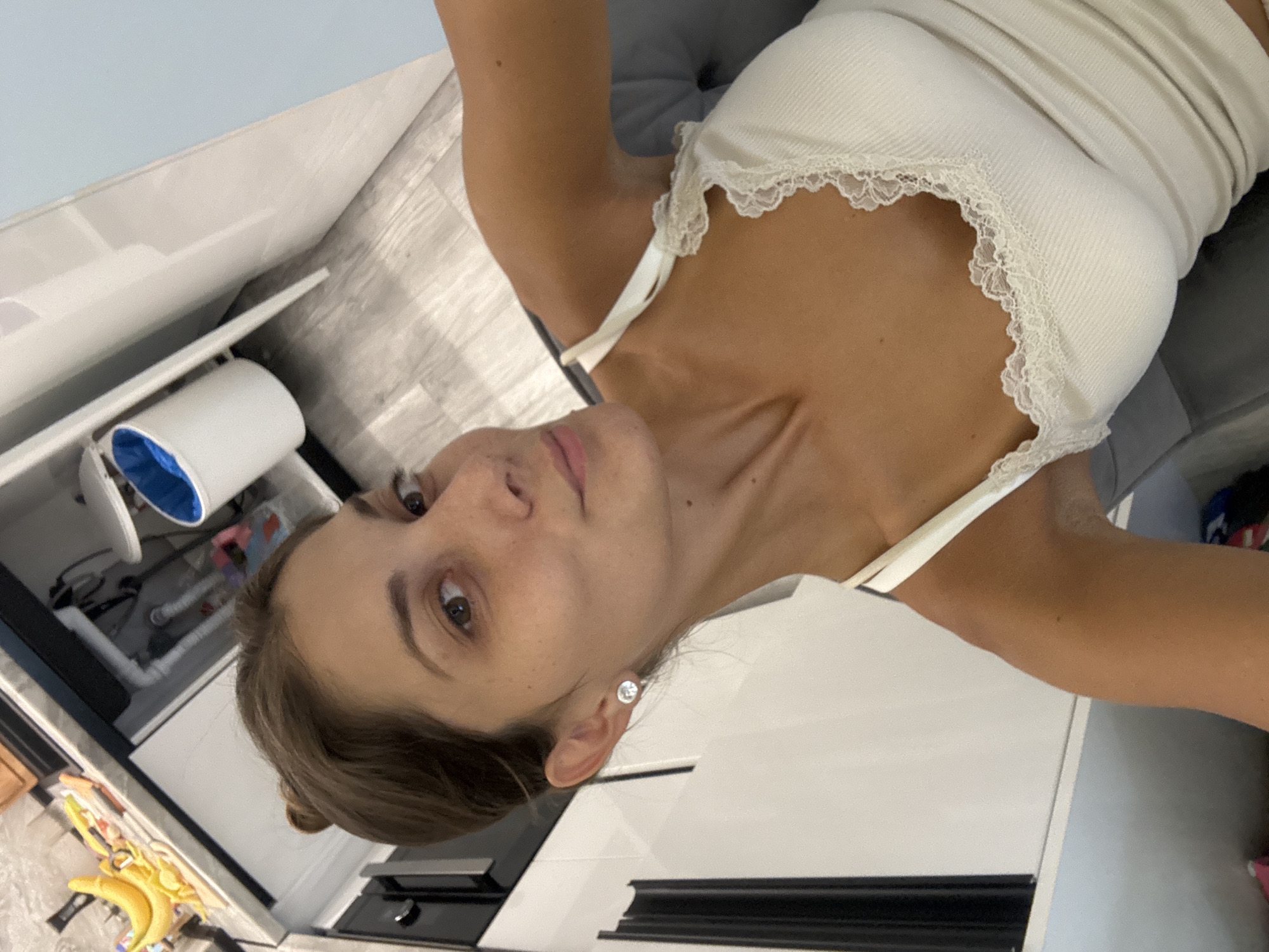 A young woman is looking at the camera. She is wearing a white top with lace trim. She is located in Burgas, Bulgaria, presumably in an apartment. The background includes kitchen elements such as white cabinets and a refrigerator containing food items, including bananas.