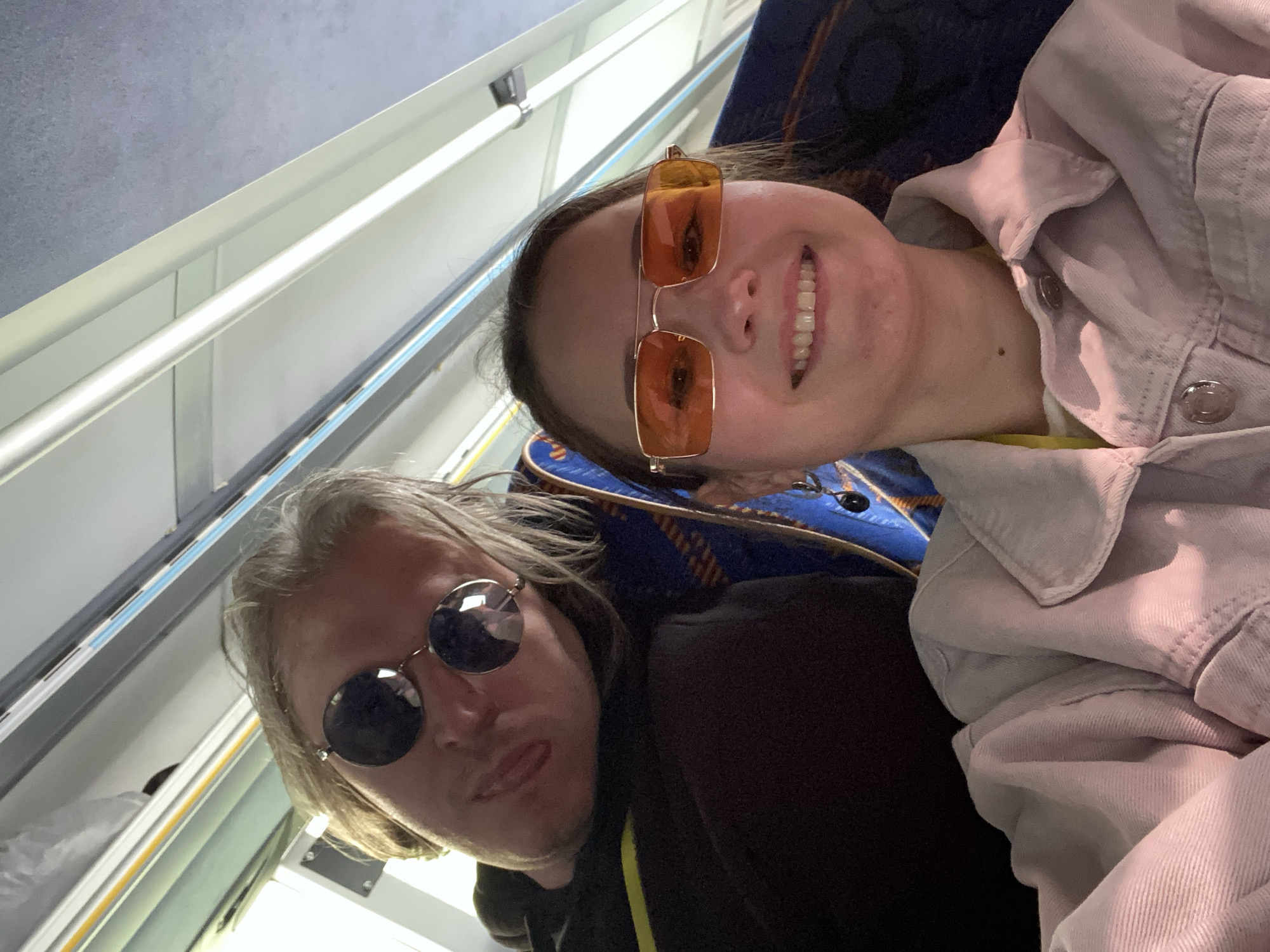 Two people are posing for a selfie. The person on the right is wearing orange sunglasses and a light-colored jacket. The person on the left has blond hair and is wearing dark sunglasses and a black hoodie with a yellow stripe. The setting appears to be the inside of a train carriage. Based on the information provided, it is not possible to confirm or deny if the location is in the city of Peresvet, Russia.