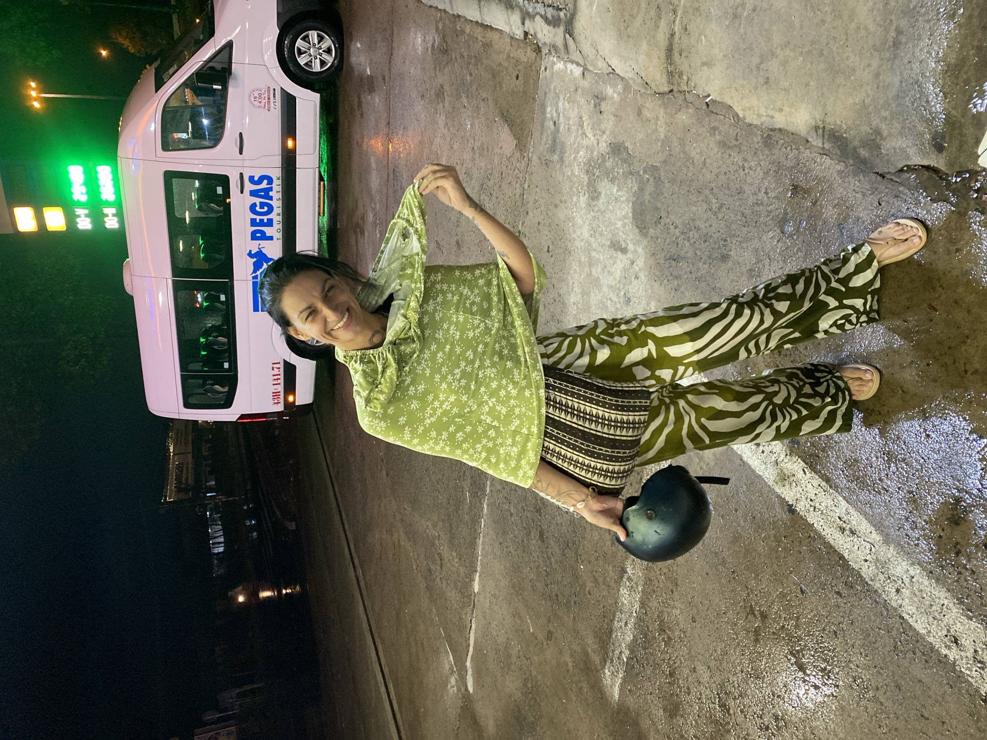 A woman with dark hair, wearing a light green floral top, green and white patterned pants, and sandals, stands smiling brightly on wet pavement. She holds up a portion of her top with her right hand and a dark motorcycle helmet with her left. The scene is illuminated by artificial lights, indicating it is nighttime.
Behind the woman is a white Mercedes-Benz Sprinter-style van. The van has "PEGAS TOURISTIK" written in blue letters on its side, along with a small circular logo containing the number "16". Further back, on the van's side, the numbers "43H 141.71" are visible in red, likely a license plate or vehicle identifier.
To the left of the woman and behind the van, a gas pump display with green illuminated numbers shows "DO-V 29400" and "DO-N 26200". The ground is damp and reflective, suggesting recent rain or wet conditions. The overall setting appears to be a roadside stop or gas station at night. The woman's cheerful expression suggests a positive mood. This scene takes place in Phu Thuy Ward, Vietnam.