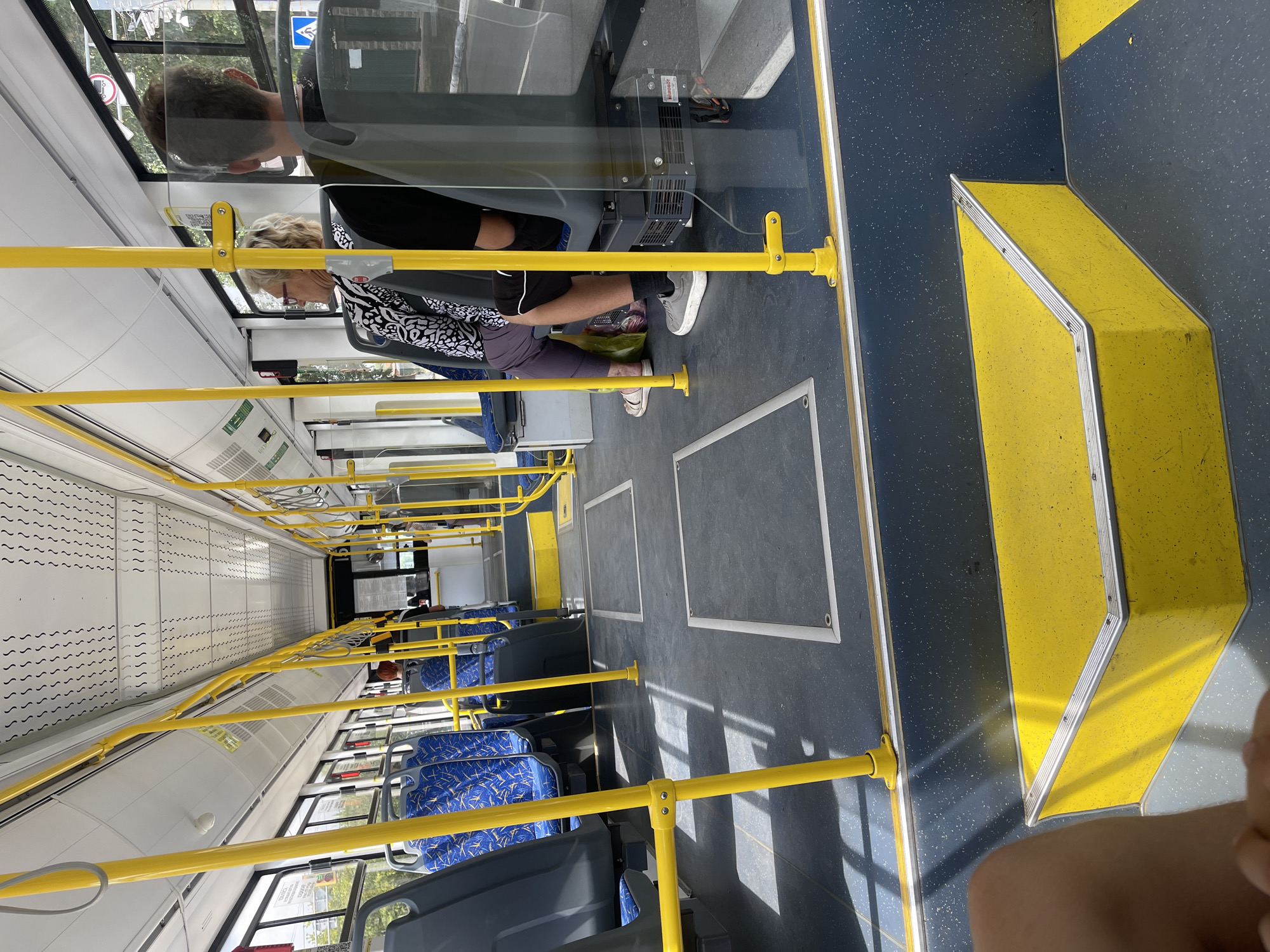 The image shows the interior of a public bus, likely in the city of Oskemen, Kazakhstan, due to its characteristic urban public transport design. The perspective is from a seated passenger looking forward. Bright yellow poles and handrails are prominent throughout the bus, a common safety feature. The seats are blue with a patterned design and are arranged in rows. Several passengers are visible, including a woman with blonde hair wearing glasses and a patterned shirt, and a man with dark hair wearing a black shirt. The floor is a dark grey speckled material. Sunlight streams through the windows, casting shadows on the floor and seats. The overall environment suggests a typical day on public transportation.