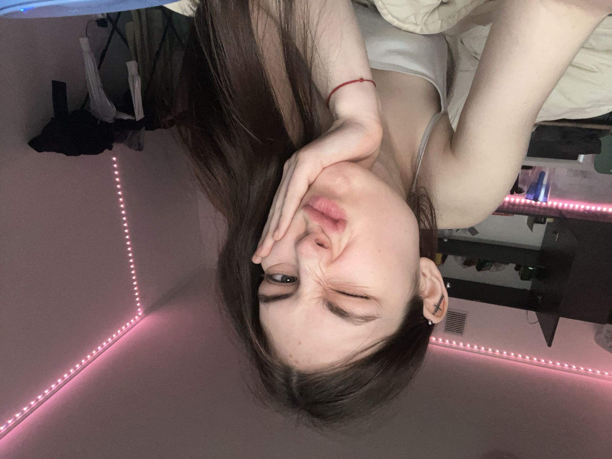 A young woman is depicted in a selfie-style photograph, taken from a close-up, low-angle perspective. She is lying down, with her head tilted to the right. She is winking with her left eye and has her hand partially covering her face, slightly pulling it. She is wearing a light-colored tank top and a red string bracelet on her left wrist. Her long brown hair is loose. The background shows a room with pink LED strip lights illuminating the ceiling and wall intersection. In the upper left, there are clothing items hanging on a rack. In the background, there are various objects on a desk and shelf, including bottles and containers. The location is indeterminate; there is no evidence to suggest that it is in Saint Petersburg, Russia.