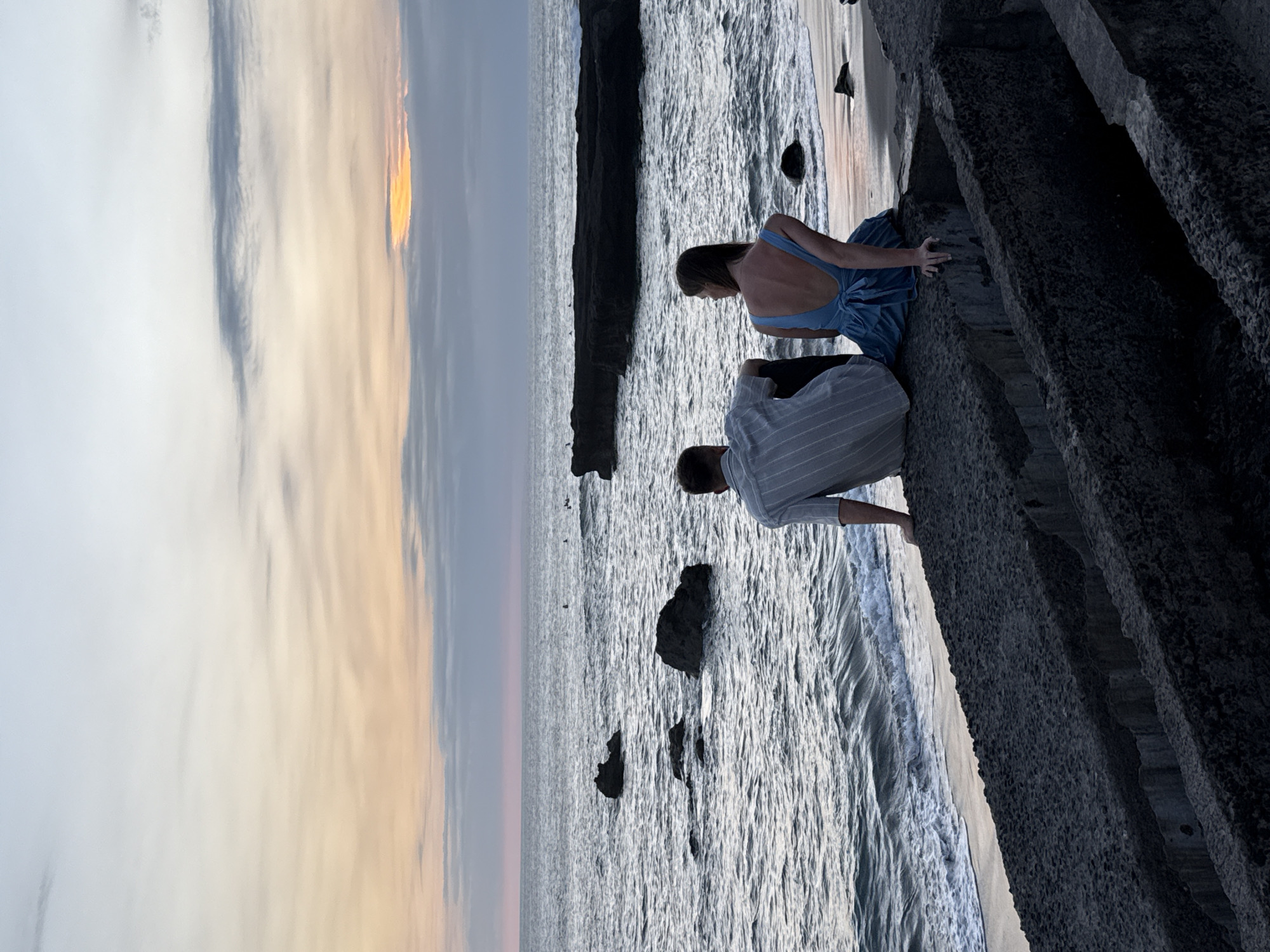 A photograph shows a couple sitting on a concrete structure overlooking the ocean at sunset. The sky is a gradient of yellow, orange, and gray. A woman in a blue dress sits to the left of the frame. A man in a light gray striped shirt is next to her. They are both looking out at the ocean. Several large, dark rocks are in the ocean. The location is North Kuta, Indonesia.