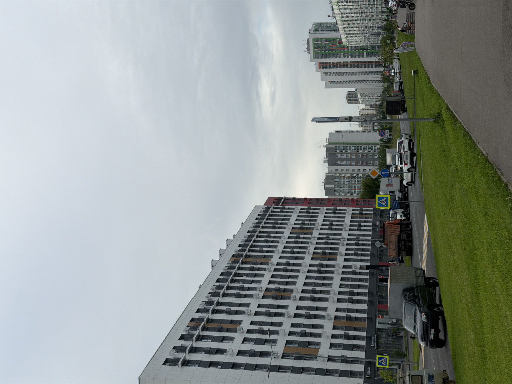 The image depicts a street scene in Kazan, Russia, characterized by modern, high-rise residential buildings. A long, white, multi-story building with numerous windows and balconies dominates the foreground. To the right of this building, a bright green grassy area runs alongside a paved pathway. Beyond the main building, other tall residential structures, some with colorful facades of green and orange, are visible in the background. The sky is overcast, suggesting a cloudy or possibly rainy day.
Several vehicles are present on the road, including a truck with a beige cargo container, a black SUV with a license plate visible, and other cars parked or driving. Pedestrian crossing signs are also visible. The overall impression is of a contemporary urban environment, likely a residential district, during daytime. The lighting is diffused due to the cloudy weather, and there are no strong shadows. The scene appears calm, with no specific events or activities immediately apparent, other than the presence of vehicles and buildings.
The text visible in the image includes what appears to be a sign with Russian text that says "СЕРВИС" and another sign with a street name or building identifier which is not clearly legible. The license plate on the black SUV is partially visible, showing Cyrillic characters and numbers.
