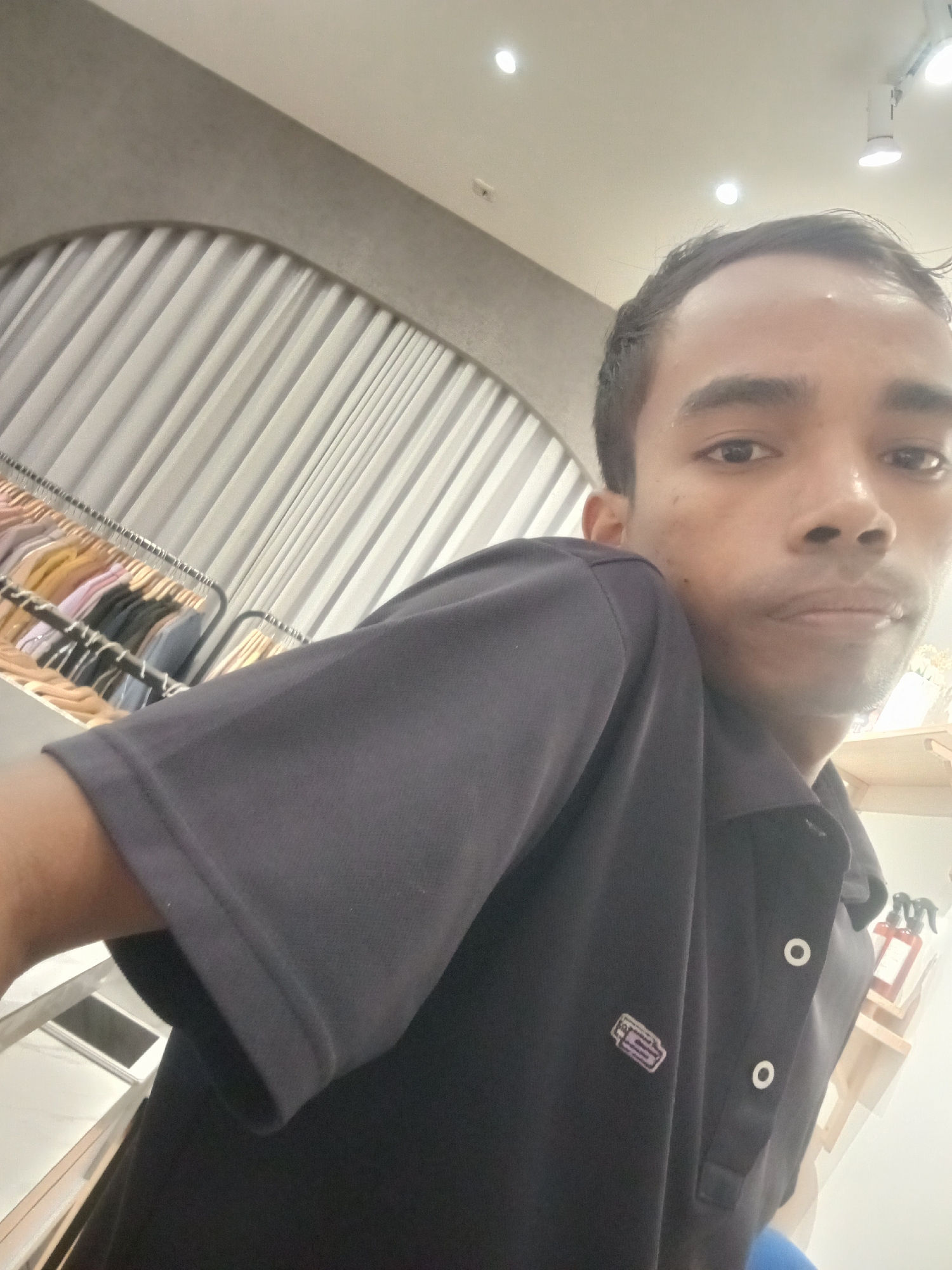 In the city of Larangan, Indonesia, a young man of South Asian descent is captured in a close-up, selfie-style photograph taken indoors. He is wearing a dark, short-sleeved polo shirt. The background is blurred but suggests an indoor environment, possibly a clothing store, with a rack of colorful shirts on hangers visible on the left. The lighting is bright, illuminating the scene and casting a slight sheen on the man's skin.