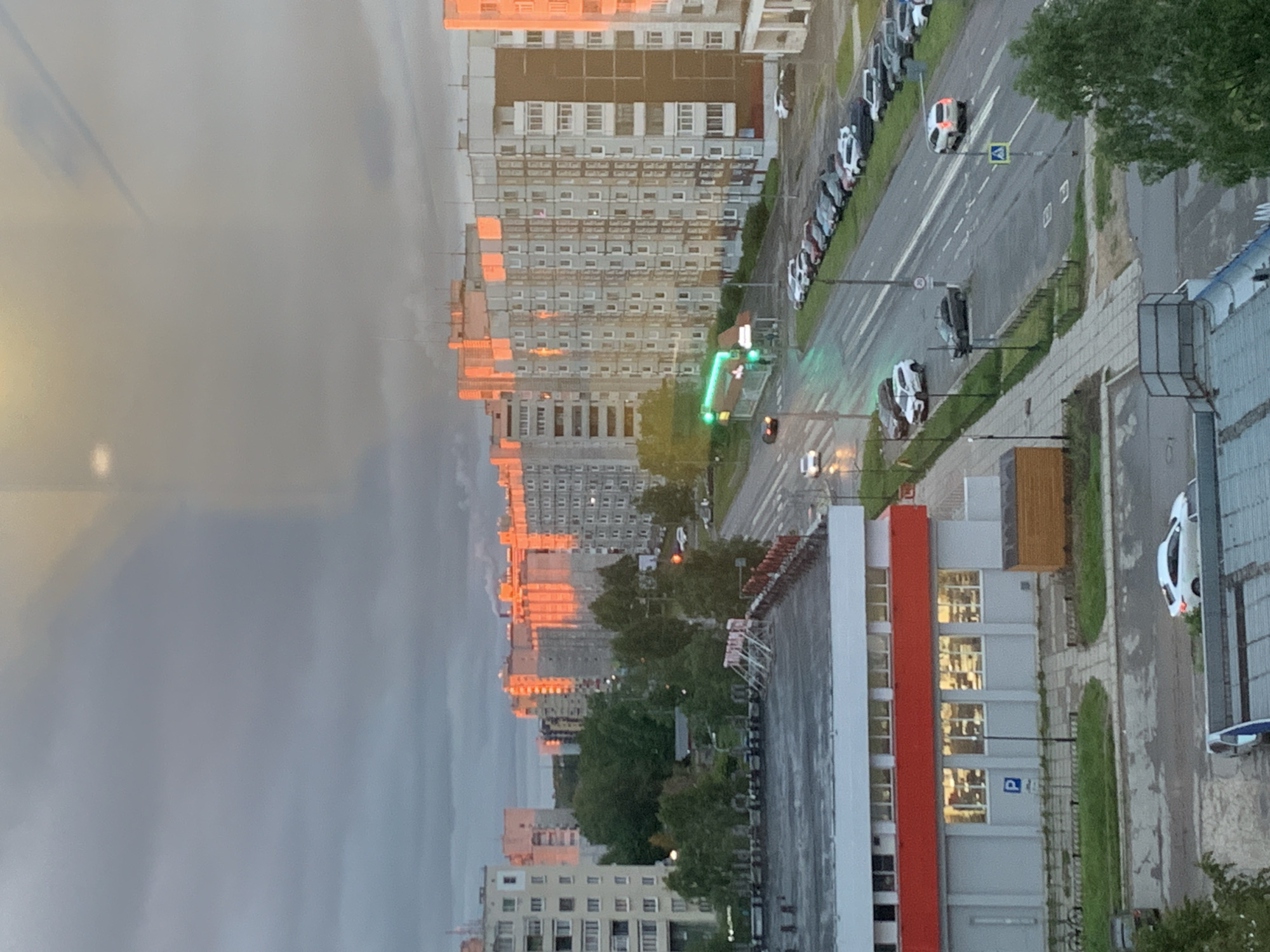 The image captures an urban street scene from an elevated viewpoint, likely late afternoon or early evening, given the low sun casting a golden-orange light on distant buildings against a grey, overcast sky. The overall atmosphere is calm and typical of an urban environment.
In the foreground, a modern building with a distinctive vertical red panel and numerous windows, some illuminated, is visible on the right. A blue "P" parking sign is affixed to this building, and a white "P" is painted on the asphalt nearby.
The central part of the image features a multi-lane road that appears wet, suggesting recent rain. White lane markings and a pedestrian crossing are visible. Several cars are parked along the left side of the road, including multiple white vehicles that resemble police cars, distinguished by roof-mounted light bars. Other cars are seen driving on the road, one with illuminated taillights. A green traffic light is clearly visible, along with streetlights and a pedestrian crossing sign.
Beyond the road, numerous multi-story residential buildings with panel-style architecture extend into the distance. The low sun dramatically illuminates their facades and windows with a warm, reflective glow. Green trees are interspersed along the road and between the buildings, adding natural elements to the concrete landscape. No people are visible.
The architectural style of the buildings, the type of vehicles, and the road infrastructure are consistent with the provided location context of Severodvinsk, Russia.