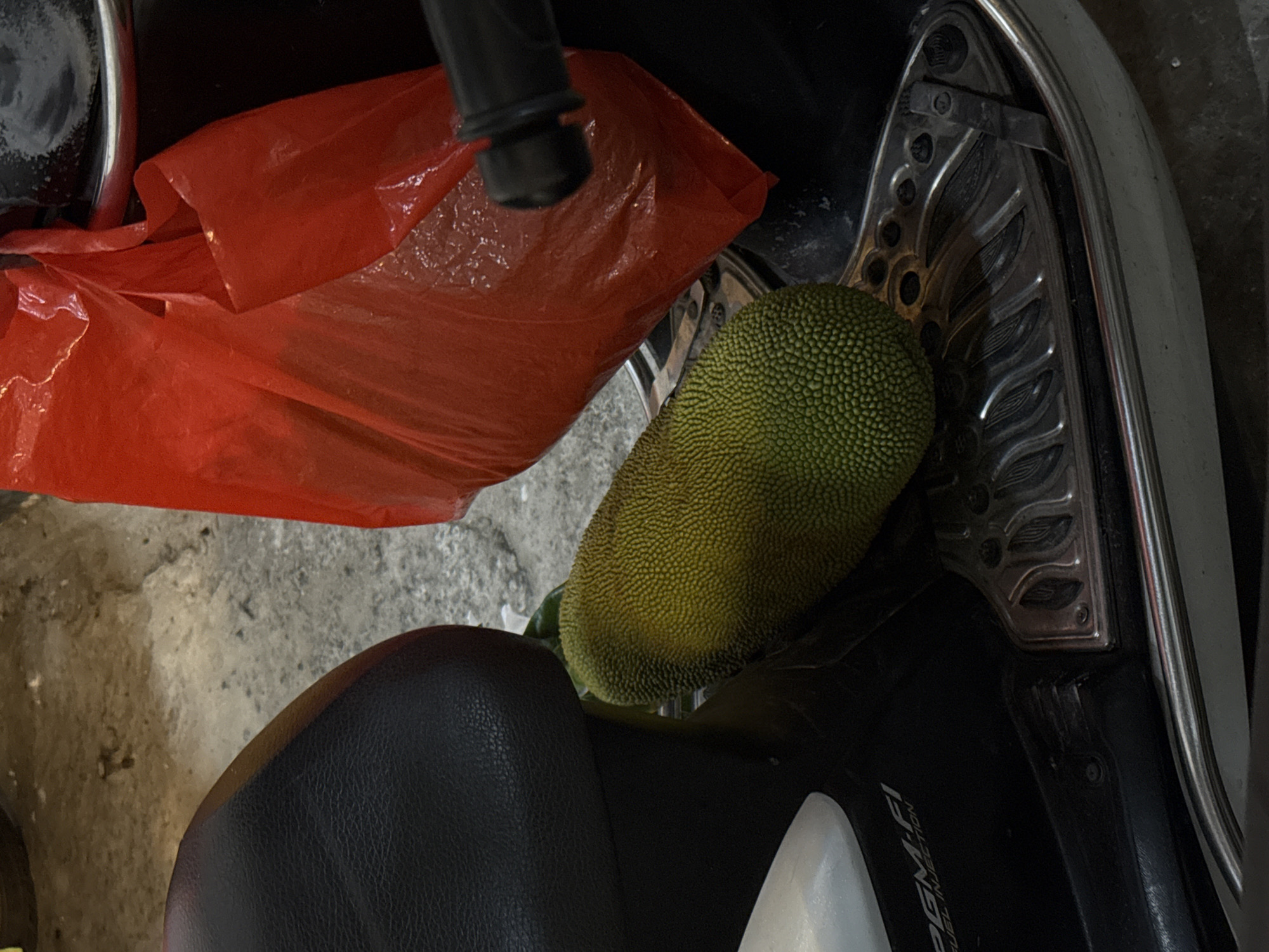 A jackfruit rests on the floorboard of a motorcycle. The jackfruit is oval-shaped and green with a spiky exterior. To the right of the jackfruit, a red plastic bag is partially visible. The motorcycle seat, which is black and textured, is to the left of the jackfruit. The ground beneath the motorcycle appears to be concrete. The scene is dimly lit, suggesting it is either nighttime or indoors.