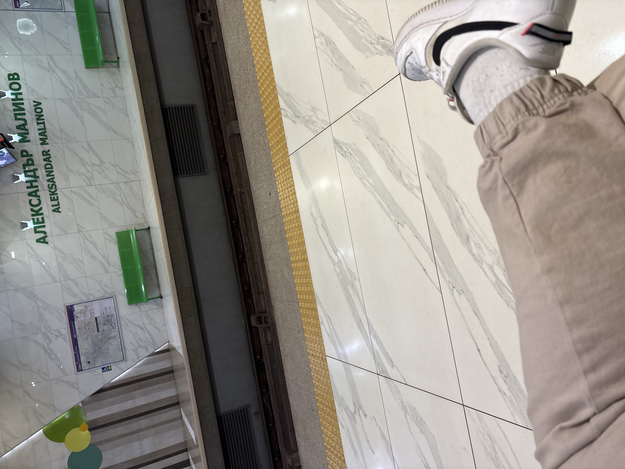 The image depicts the Aleksandър Malinov metro station in Sofia, Bulgaria. The station platform features tiled flooring, tactile paving along the edge, and seating. A ventilation grate is visible on the wall. A person's leg, wearing a white Nike shoe, grey sock, and beige trousers, is positioned on the right side of the image.