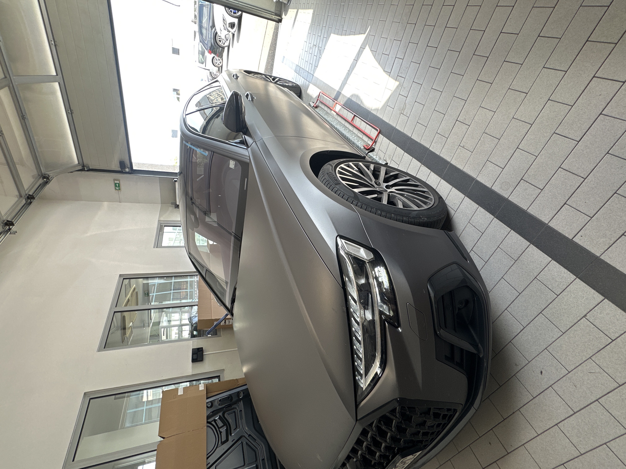 An Audi A8 sedan, finished in a matte grey wrap, is parked inside a garage with a large open overhead door. The car is positioned at an angle, facing towards the left of the frame. Its front end, including the distinctive grille and headlights, is clearly visible. The vehicle has custom wheels and a sporty bumper. To the left of the car, a car hood is propped up and leaning against some brown cardboard boxes. In the background, through the open garage door, several other cars are parked outside. The garage floor is tiled with a light grey and white pattern, with a dark grey strip running diagonally across it. Sunlight streams into the garage, casting shadows on the floor. The walls of the garage are painted white.