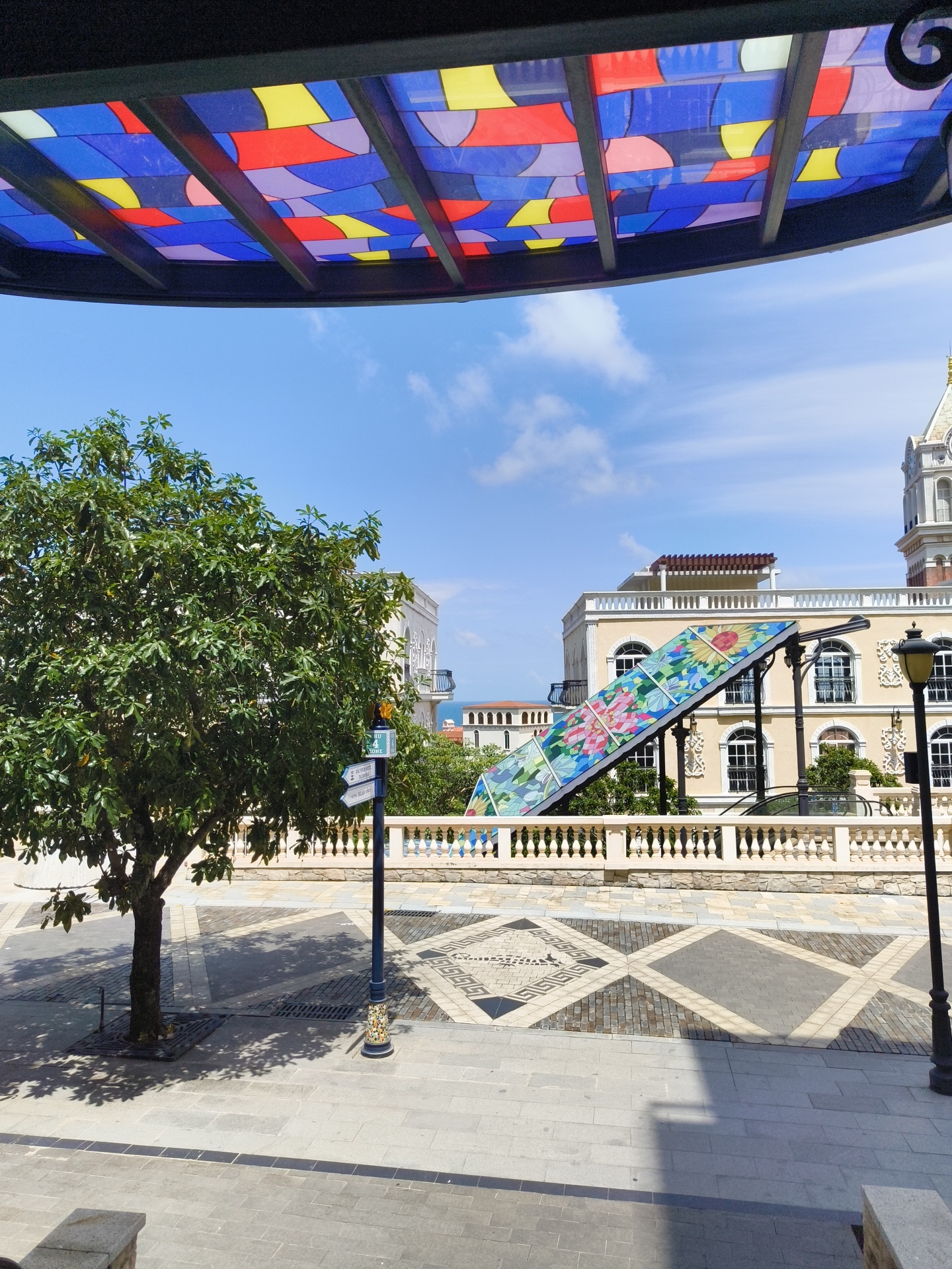 The image shows an outdoor scene in a city, specifically Phú Quốc, Vietnam. In the foreground, a paved walkway with a decorative pattern leads towards buildings in the background. A large tree with green foliage stands to the left of the walkway. A streetlamp is positioned on the right side of the walkway. Further back, a decorative, colorful glass-covered ramp or walkway with a floral design is visible, leading up to a light-colored building with arched windows. Another building, appearing to be a clock tower, is partially visible on the far right. The sky is blue with scattered white clouds.