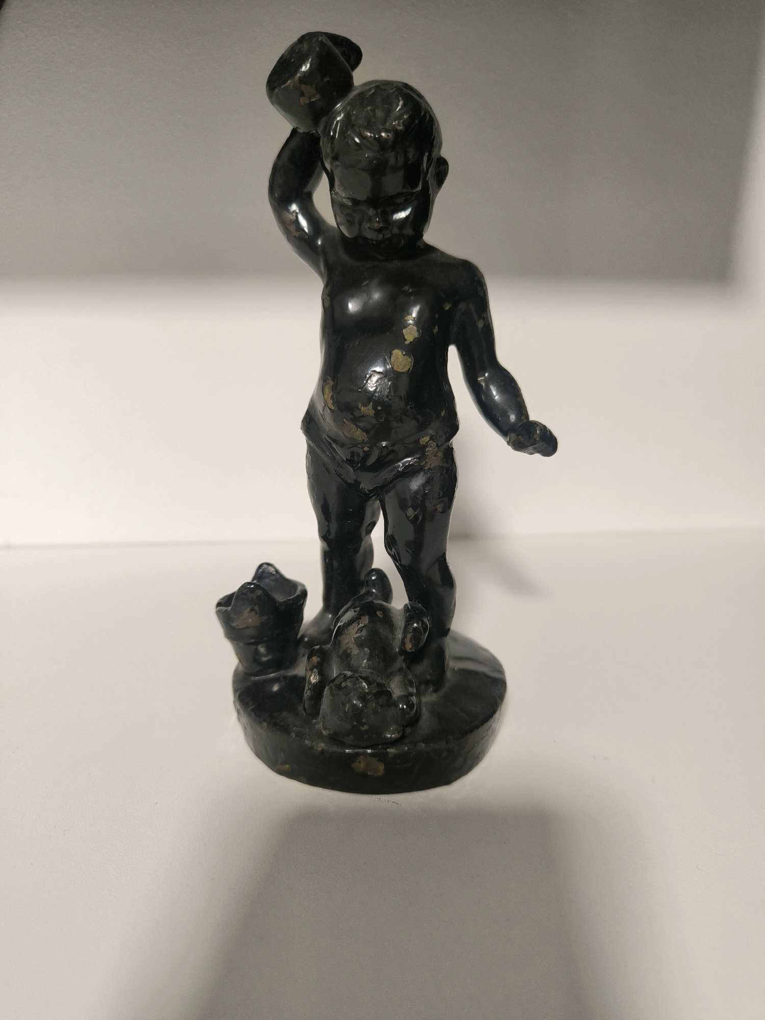 This is a close-up, eye-level photograph of a dark, possibly bronze, figurine of a baby or cherub standing on a circular base. The figurine is shiny and shows some wear, with patches of lighter color, possibly brass or gold, showing through the dark patina. The cherub is depicted in a dynamic pose, with its right arm raised, holding a small object that is difficult to identify, possibly a cup or a jug. Its left arm is extended slightly forward. The cherub is unclothed, with stylized anatomical features.
At the base of the figurine, nestled around its feet, are two small, dark, abstract shapes. One appears to be a small animal, perhaps a dog, lying down, while the other is less distinct, possibly a rolled-up cloth or another object. The figurine is placed on a flat, white surface, which could be a shelf or a table. The background is a softly lit, neutral gray, creating a shallow depth of field that keeps the focus on the figurine. The lighting casts a soft shadow of the figurine onto the white surface. There is no visible text in the image. The scene suggests a decorative object, likely an antique or a collectible, presented in a display setting.