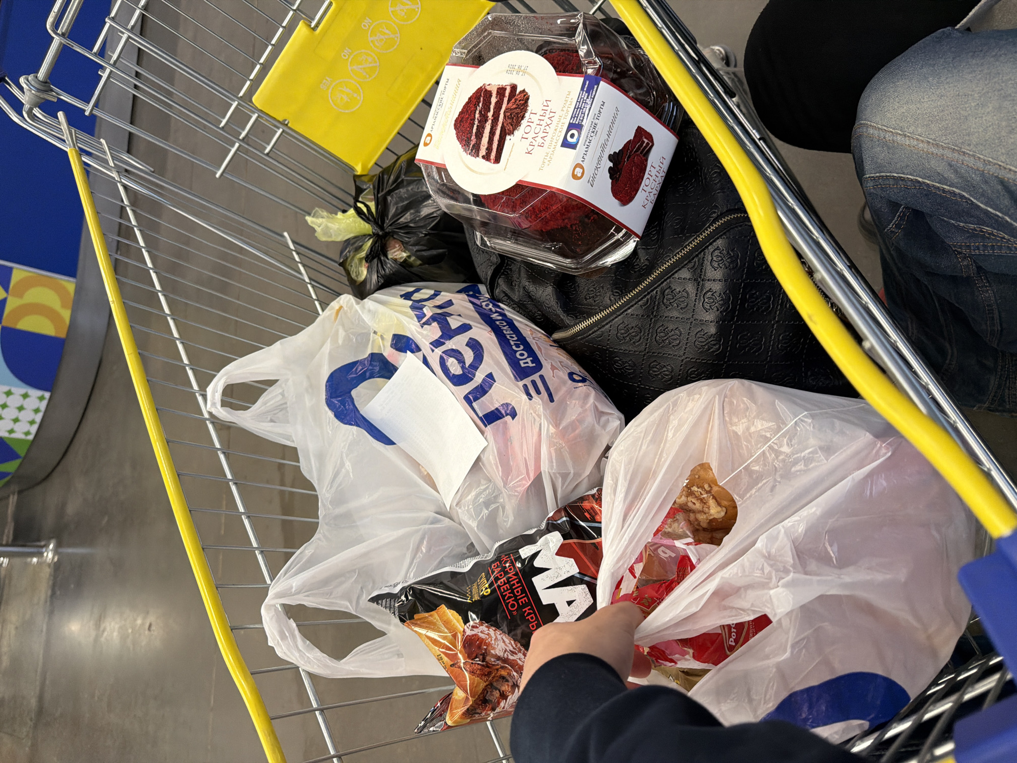The image shows a close-up view of a shopping cart filled with groceries and other items. A person's hand is visible, reaching into a white plastic bag. The cart contains several plastic bags, one of which holds a red velvet cake in a clear plastic container. Another bag has a label with Cyrillic text and an image of food. A black bag is also present. The shopping cart itself is made of metal and has a yellow bar along the top. The background appears to be a store aisle, with a polished concrete floor and some shelving or displays visible in the distance. The scene is indoors, likely a supermarket or grocery store, and it is plausible this is in Nizhny Novgorod, Russia, given the presence of Russian language on product packaging.