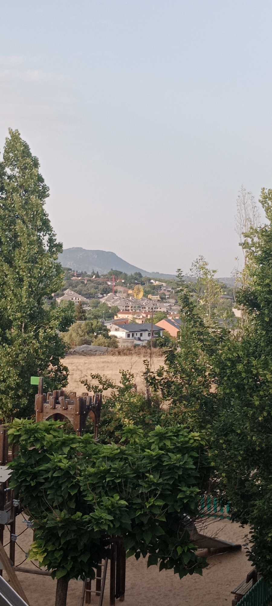 The image captures a scenic view overlooking the town of Galapagar, Spain, from a slightly elevated vantage point. In the foreground, a children's playground structure is visible, featuring wooden elements resembling a castle, complete with a small green flag atop one of its turrets. Lush green foliage from trees and bushes partially obscures the playground and the middle ground.
Beyond the playground, a dry, golden field stretches out, dotted with a few trees and rocks. In the mid-distance, the town of Galapagar unfolds, characterized by a cluster of buildings with terracotta roofs and white or light-colored walls. Notably, a large, golden, spoked structure, possibly a decorative element or part of a larger installation, stands out among the buildings. A red crane is also visible in the town.
The background is dominated by a hazy, pale blue sky and a range of distant hills or mountains, softened by atmospheric conditions. The overall impression is a tranquil, late afternoon or early evening scene in a semi-rural setting, with elements of recreation and residential life combined with natural landscapes. There are no people visible in the image.