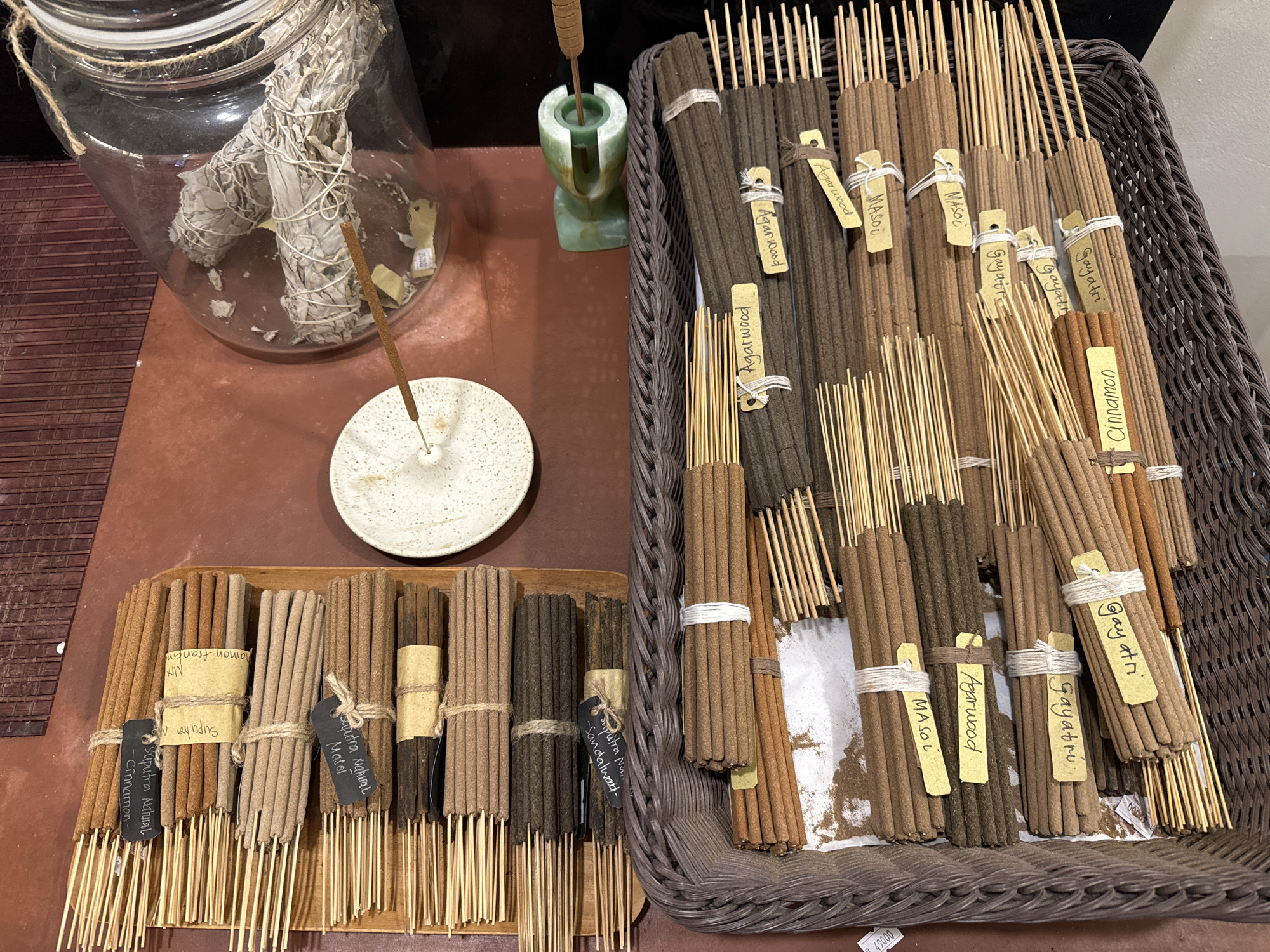 The image shows a collection of incense sticks displayed for sale. In the upper left, a large glass jar contains several bundles of dried sage, commonly used for smudging. An incense stick is lit and burning, resting on a small, speckled ceramic dish. To the right of the jar, a green jade-like incense holder holds the lit stick.
The majority of the image is filled with various bundles of incense sticks arranged in two main areas. The left side features incense sticks laid out on a wooden tray, organized into several rows. Each bundle is tied with twine, and some have small tags or labels identifying the scent, such as "Suputra Natural - Cinnamon-" and "Suputra Natural - Sandalwood-". The sticks vary in color from light brown to dark brown.
On the right side, a large, woven basket overflows with more bundles of incense sticks, many of which are darker in color. These bundles are also tied with twine and have yellow tags with handwritten names like "Agarwood", "MASOL", and "Gayatri". The incense sticks are predominantly in shades of brown, with some lighter wooden sticks visible among them. The overall presentation suggests a market or shop setting where these aromatic products are offered for sale. The location is identified as Mengwi, Indonesia.