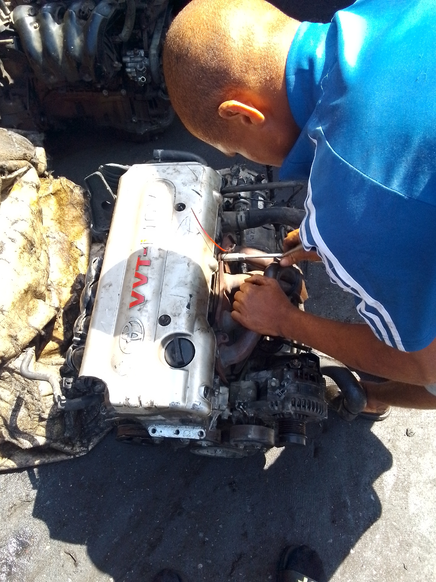 The image shows a man working on a car engine outdoors. The man, wearing a blue shirt with white stripes on the sleeves, is hunched over the engine, manipulating parts with his hands and a tool. The engine is a Toyota VVT-i, lying on the ground next to a stained cloth. In the background is another engine. The scene is taking place on a concrete surface, likely in an auto repair environment. Given the reference, the location is inferred to be in Onitsha, Nigeria.