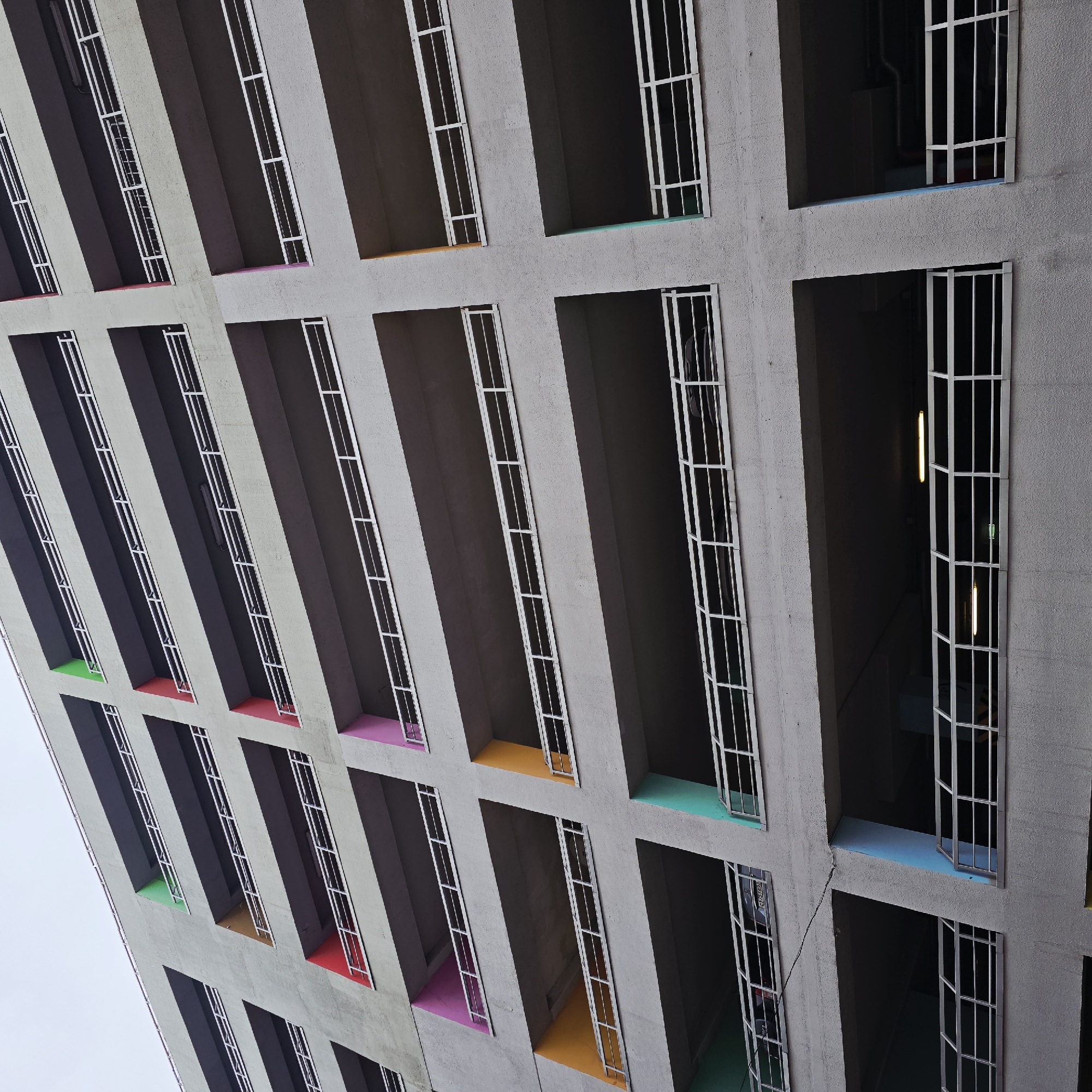 The image captures a low-angle view of the open-air stairwell balconies of the House of the Commune building in Moscow, Russia. The building features a brutalist architectural style with recessed balconies and white metal guardrails. The balconies are painted in different colors, including pink, green, orange, and blue. The walls of the building are gray. There is sky visible in the background. In one of the balconies, an open door and interior lights can be seen.