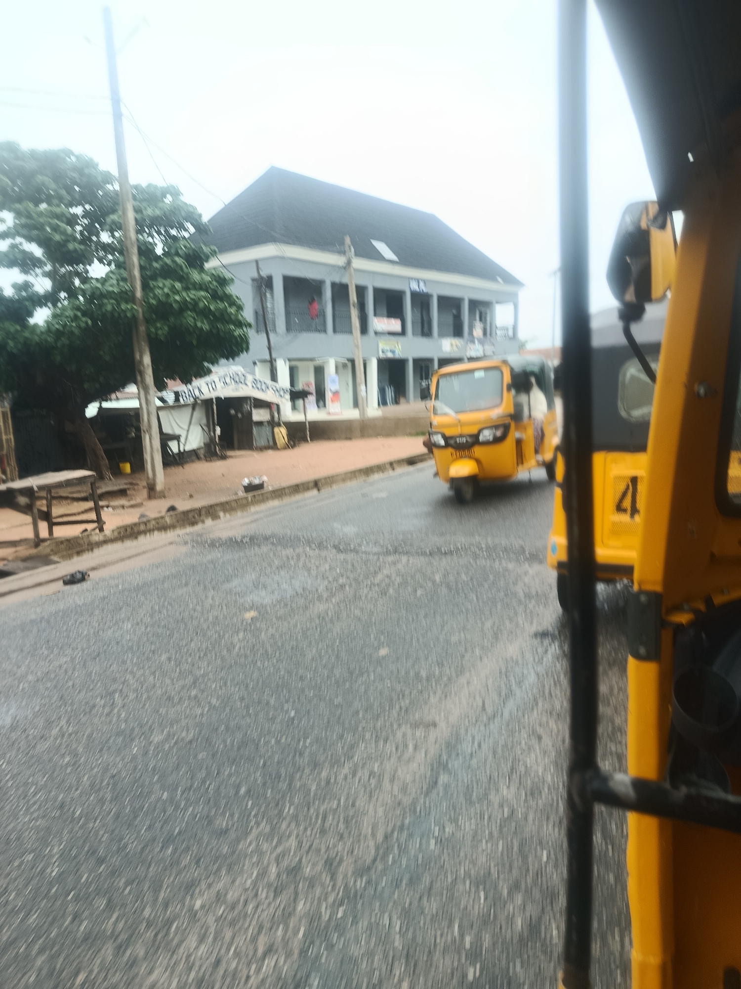 The image shows a street scene in Jalingo, Nigeria. In the foreground, the bright yellow body of a tuk-tuk, also known as an "okada" or "motorcycle taxi," dominates the right side of the frame. Part of its black roof and a black metal bar are also visible. Moving away from the viewer, the road surface is visible, appearing wet and textured. Further down the road, another yellow tuk-tuk is traveling, with a person visible in the driver's seat and another passenger behind them. To the left of the road, there is a sidewalk area with a small, temporary structure that appears to be a shop or stall, marked with "BACK TO SCHOOL BOOKSHOP." Behind this stall, a large tree with dense green foliage stands next to a utility pole with wires. Beyond the tree and stall, a two-story building with a dark, sloped roof and grey exterior is visible. The sky is overcast and appears bright, suggesting a cloudy or hazy day. The overall impression is of a typical urban street scene in a Nigerian city, with local transportation and small businesses.
