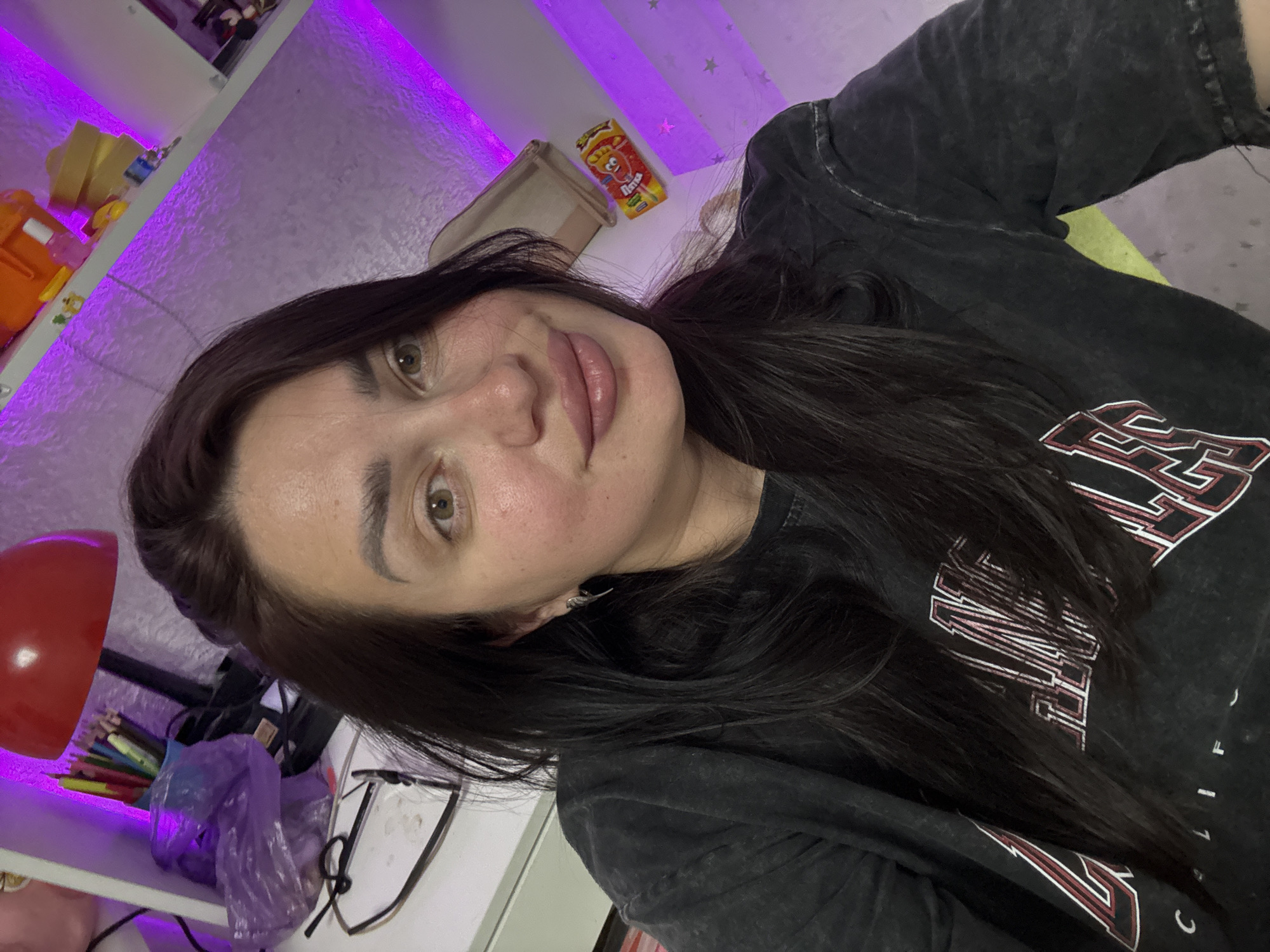 This is a selfie taken indoors, likely in a residential setting, possibly a bedroom or study, given the presence of shelves with various items and a desk with a lamp. The woman in the photo has dark brown hair and is wearing a dark t-shirt. She appears to be looking directly at the camera. The background features shelves illuminated with purple LED lighting, displaying objects such as pencils, a red lamp, plastic bags, and a packaged snack. The overall ambiance suggests a casual and personal space.