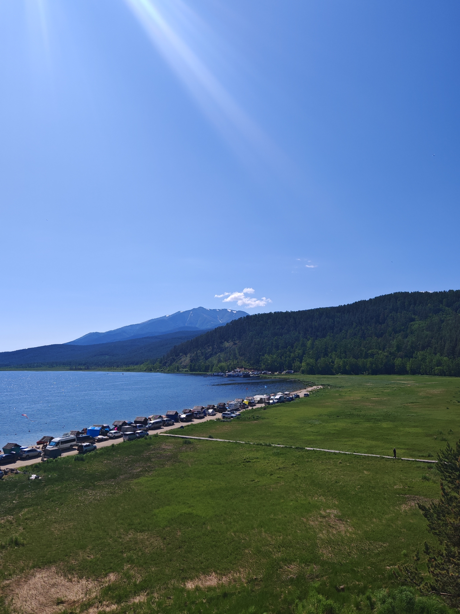 This media file captures a scenic landscape dominated by a large, calm body of water on the left and a vast, green grassy field on the right. A dense, dark green forest covers the rolling hills that rise behind the water's edge, and further in the distance, majestic mountains, some capped with snow, reach towards a clear, bright blue sky. Sunlight streams down from the upper left, creating visible rays.
Along the edge of the water, a line of vehicles and what appear to be small structures or tents are parked closely together, suggesting a gathering or settlement. The water itself is a vibrant blue, reflecting the sky. The grassy field is lush and appears well-maintained, with a single person walking along a narrow path or boardwalk that traverses the field. The time of day appears to be daytime, possibly late morning or early afternoon, given the brightness of the sunlight and the clear sky, which features a few wispy white clouds near the mountains. The overall mood conveyed is one of peacefulness and natural beauty, with a hint of human activity at the water's edge. No visible text is present in the image.