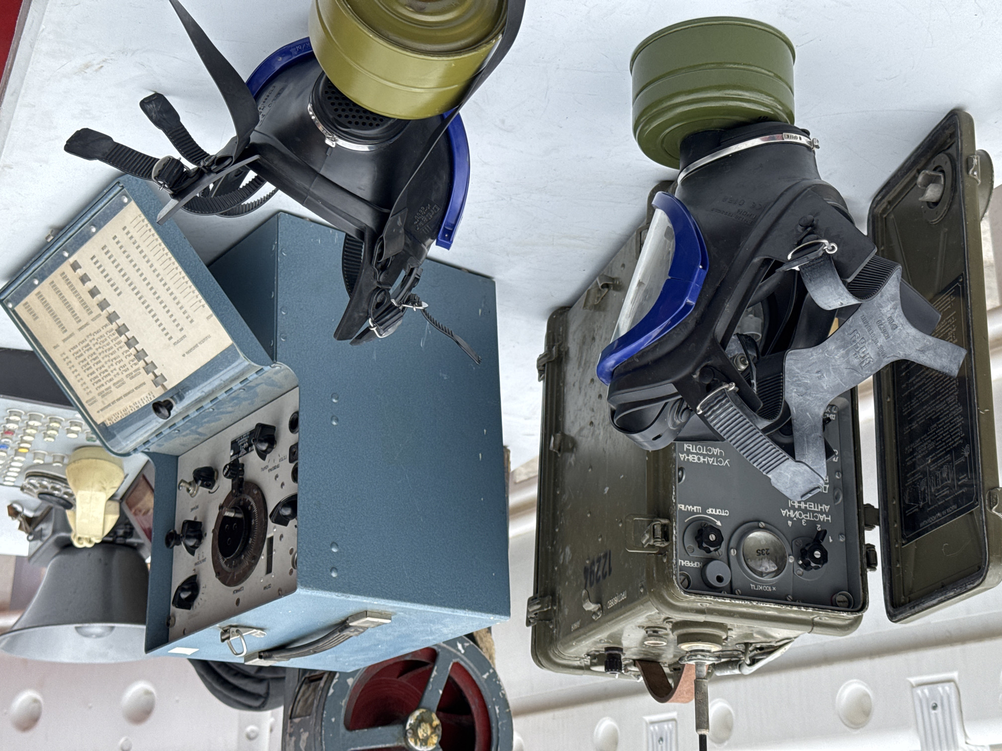 The image shows two gas masks, one with a black face piece and a yellow filter and the other with a black face piece and a green filter, alongside two military-style radio sets. The gas masks are connected to their respective radios, which appear to be of Soviet or Eastern European design. The radio on the left is blue with a control panel, while the one on the right is olive green with Russian lettering. The image is taken at an angle, and the items are placed on a white surface with circular patterns. The setting is not recognizable, and there is no information about the specific location in the city of Unknown, Unknown.