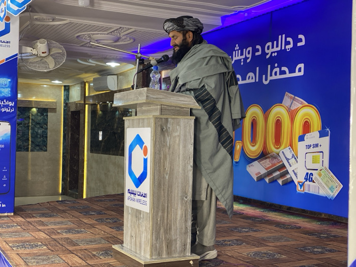 In Kandahar, Afghanistan, a man stands at a wooden podium, speaking into a microphone. He wears a black turban and a grey tunic with dark patterned stripes. A clear plastic water bottle is positioned on the podium, which features the blue and white "Afghan Wireless" logo and text in both English and Dari/Pashto. Behind the man, a large blue banner displays "Afghan Wireless" branding. The left side of the banner shows part of a smartphone image with the number "330" and "4G" text. The right side of the banner contains large white/gold Dari/Pashto text (which appears to be "د چا ليو و وبش محفل اهد"), prominent gold numerals ending in "000," stacks of banknotes, and mobile SIM cards labeled "TOP SIM +" and "4G." The indoor environment includes a white, ornate ceiling with recessed lighting. The walls are adorned with dark green/black marble-patterned panels featuring vertical illuminated gold strips. The floor is covered with a brown, geometric-patterned carpet. A white wall-mounted oscillating fan and a white wall-mounted air conditioning unit are visible in the upper background.