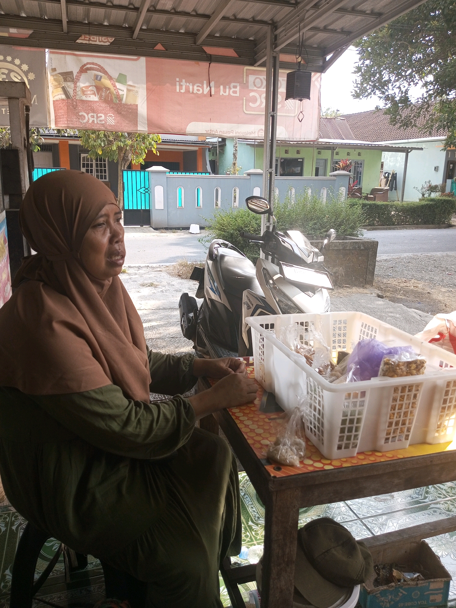 In Kalikidang, Indonesia, a middle-aged Indonesian woman wearing a brown hijab and a green dress sits at a wooden table outdoors, facing left, and looking towards something out of frame. Her hands are resting on the table near a white plastic basket filled with various items, including small bags of snacks, a purple bag, and what appears to be nuts or dried food. To her left, a portion of a dark blue chair is visible. Behind her, a motorcycle is parked. The table is set up under a roofed structure with a metal ceiling. In the background, a street is visible with houses and trees. A banner with text and an image of a basket of goods hangs on the wall behind the woman.