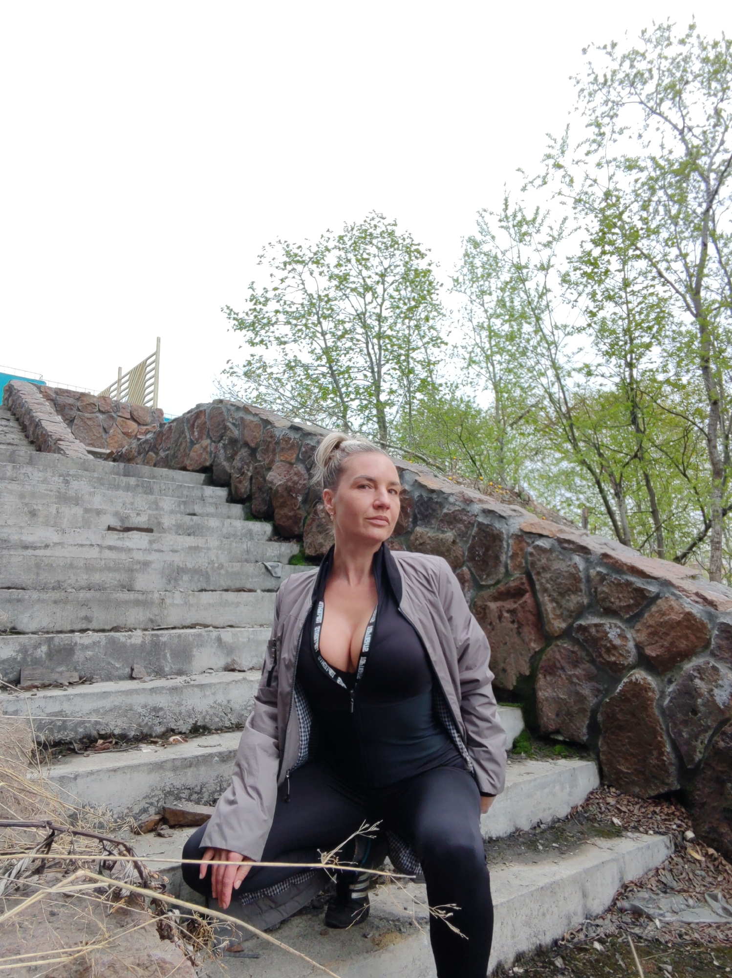 In Petropavlovsk-Kamchatsky, Russia, a Caucasian woman with blonde hair pulled back in a ponytail is crouched on concrete stairs outdoors. She is wearing a grey jacket over a black athletic top that reveals cleavage, and black leggings. The stairs are flanked by a stone wall and sparse greenery, with trees visible in the background under an overcast sky. The environment appears somewhat neglected, with debris on the stairs and dry vegetation in the foreground. The woman is looking to her right.