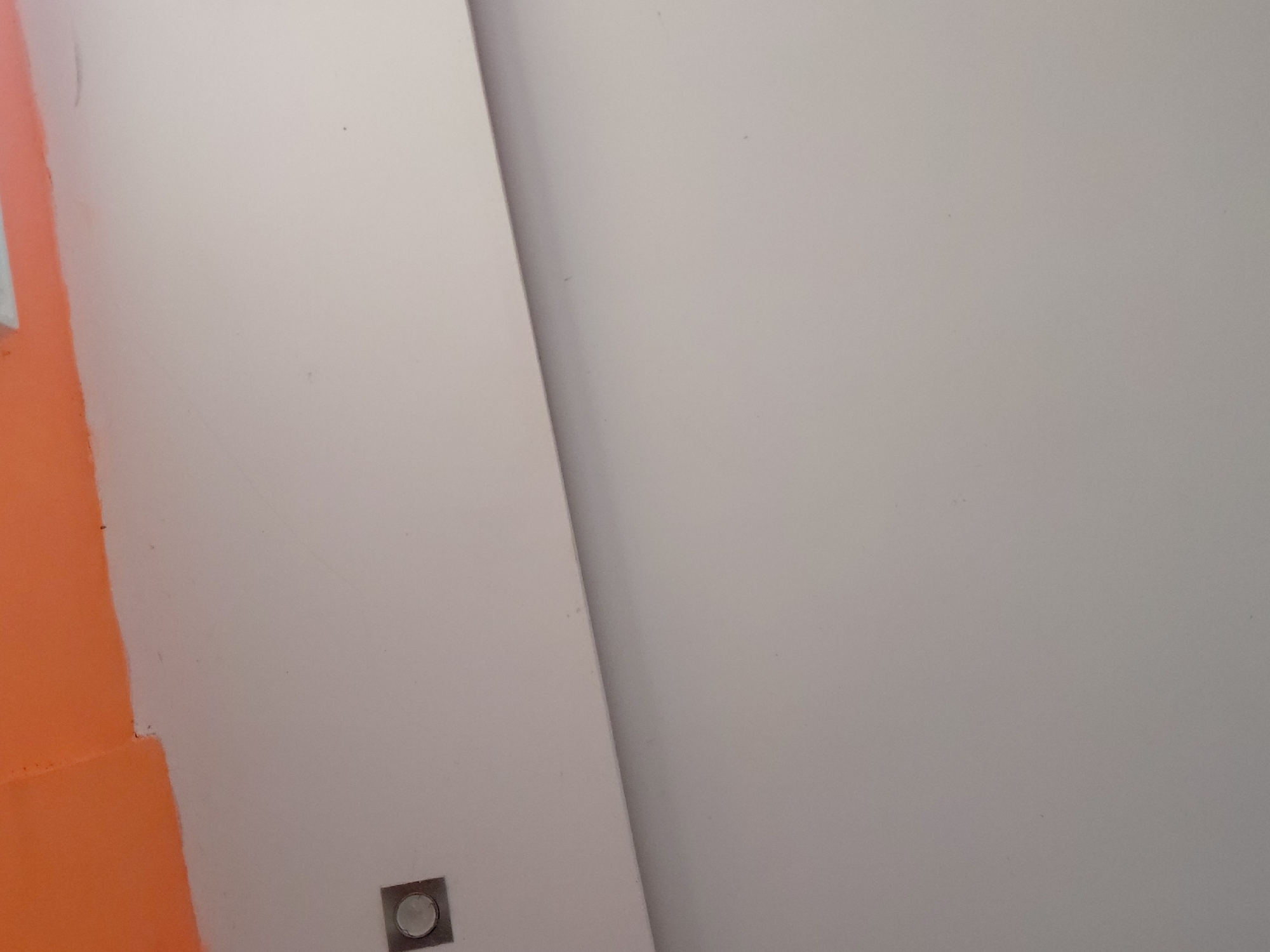 The image captures an abstract, static view of an interior space. The dominant features are two large, flat surfaces that meet at a sharp diagonal line, creating a division across the frame. The majority of the right and upper sections of the image display a smooth, light grey or off-white wall. The left side also features a light grey/off-white surface, but a significant portion of its left-hand side is painted a vibrant, solid orange, with a slightly uneven paint line.
In the bottom-left area, partially overlapping both the orange and light grey sections of the wall, there is a small, square wall fixture. This fixture appears metallic and contains a circular element in its center, possibly a button, sensor, or indicator light. No people or activities are visible. The lighting is even, typical of an indoor setting, and no specific time of day or weather conditions can be inferred. There is no visible text. This interior scene is located in Santa Ana, Venezuela.