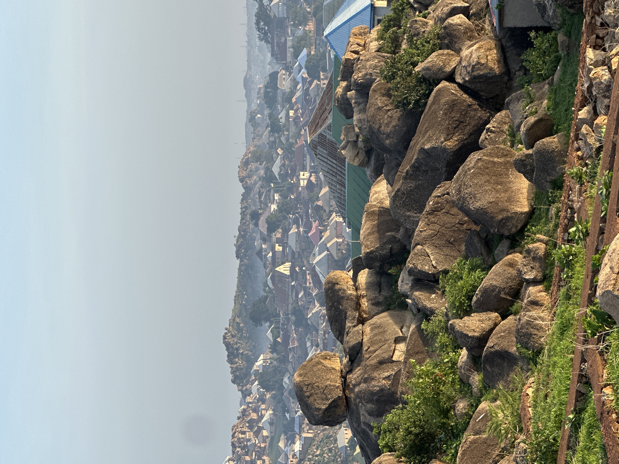 The image presents an expansive, high-angle view of a town nestled within a rugged, rocky landscape, consistent with the geography of Jos, Nigeria.
In the foreground, a substantial collection of large, irregularly shaped boulders dominates the lower and right-hand side of the frame. These rocks are primarily dark grey and brown, displaying weathered surfaces. Green shrubs and patches of grass grow between the rocks and on ledges, adding hints of vegetation. A rusted metal structure, possibly a railing or support beam, is partially visible on the far right edge, embedded amongst the rocks.
Beyond the immediate rocky outcrop, the midground and background reveal a sprawling human settlement. Numerous buildings, mostly residential, are tightly packed across undulating terrain. The rooftops exhibit a variety of muted colors, including grey, brown, red, blue, and green, suggesting different materials, with corrugated metal being common. A few larger buildings with distinct blue and teal roofs stand out closer to the rocky foreground. The distant parts of the town are enveloped in a noticeable atmospheric haze or dust, blurring details and blending into the sky.
The upper portion of the image is filled with a pale, light blue to off-white sky, indicating a bright but possibly hazy daytime. No specific activity or event is taking place, and there are no visible people or discernible text within the image. The scene offers a broad, static panorama of urban life coexisting with natural geological features under a hazy sky.