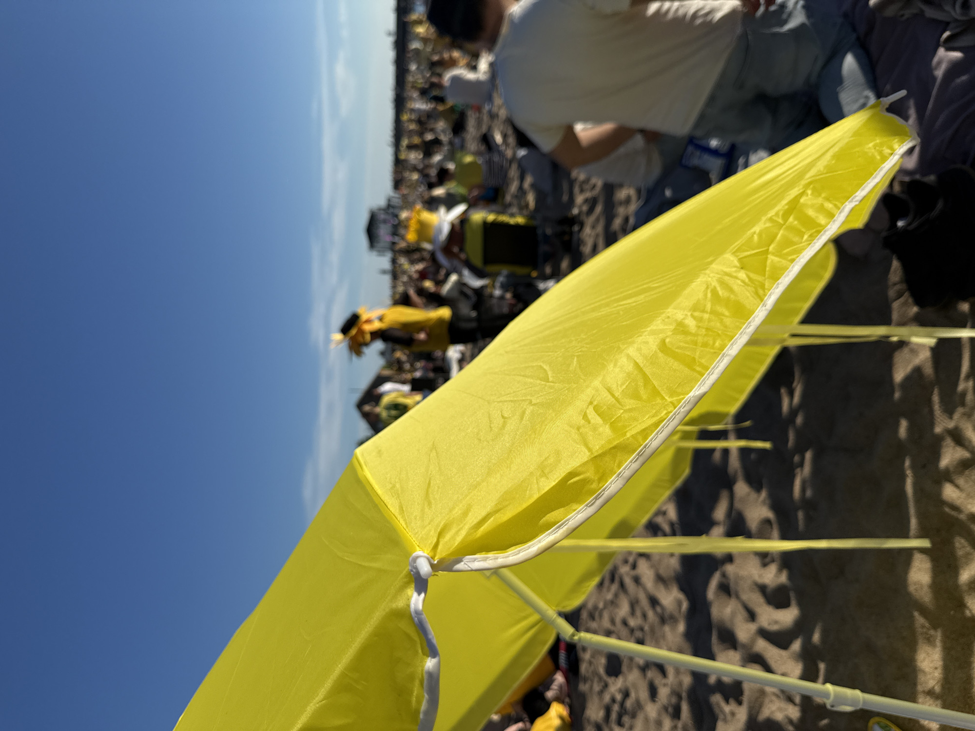 A bright yellow beach umbrella dominates the foreground of this outdoor scene, casting shadows on the sand. In the background, a crowd of people are gathered on a beach under a clear blue sky. Many attendees are wearing yellow clothing, and some have elaborate yellow headdresses, suggesting a themed event or festival. The overall atmosphere appears festive and relaxed, with people sitting and standing on the sand. The image is taken from a low angle, emphasizing the scale of the gathering and the vastness of the sky.