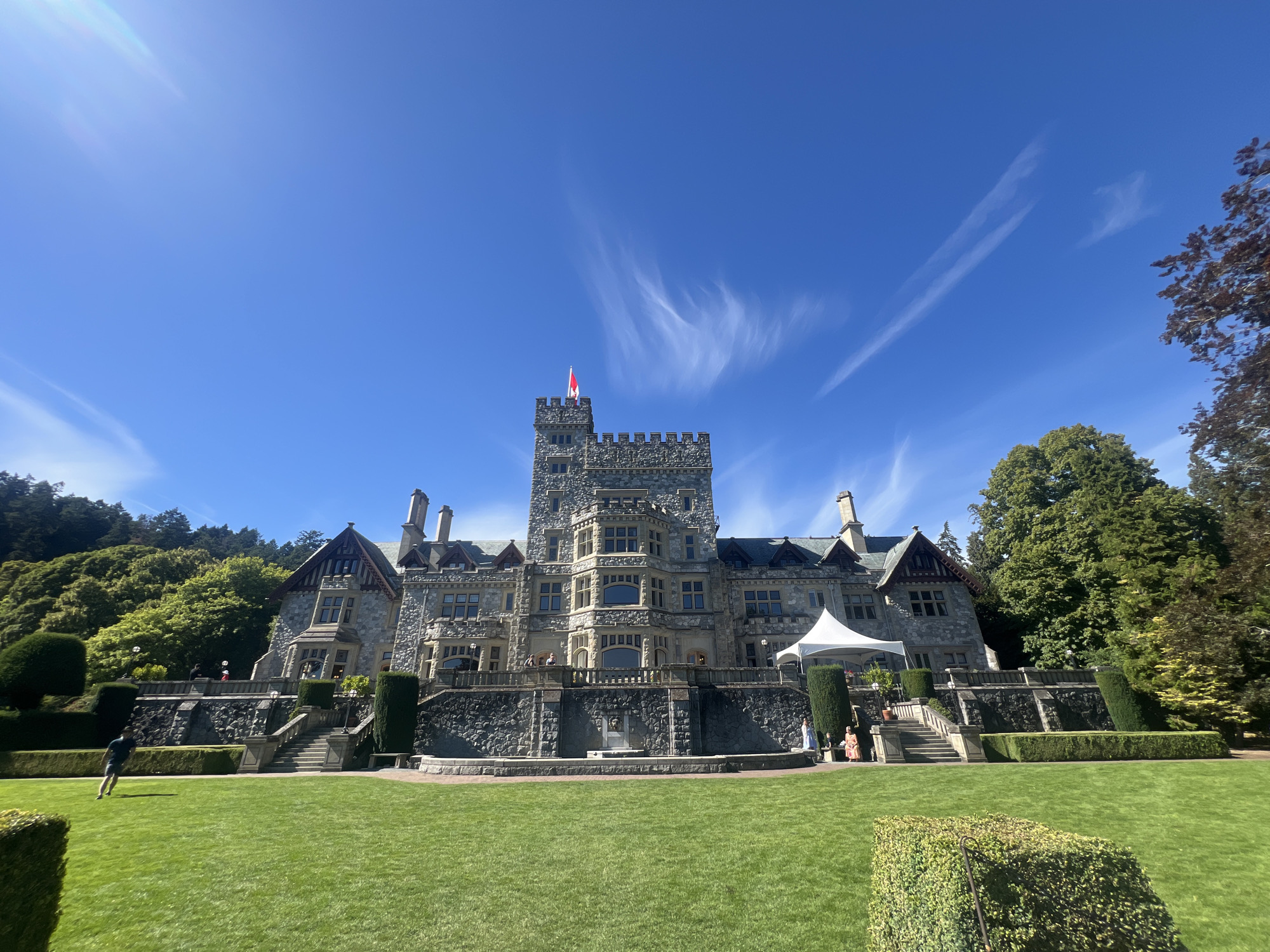 This is a full, outdoor, eye-level, wide-angle shot of a large stone castle under a bright blue sky with scattered wispy clouds. The castle, located at the Butchart Gardens in Brentwood Bay, British Columbia, Canada (near the city of Langford), features a tall central tower topped with a Canadian flag. A white tent is set up on the right side of the castle's terrace. The castle is surrounded by meticulously maintained green lawns, manicured hedges, and a stone retaining wall. Several people are visible on the castle terrace and lawn, and one person is running on the lawn in the foreground. The background consists of lush green trees and vegetation on a hillside.