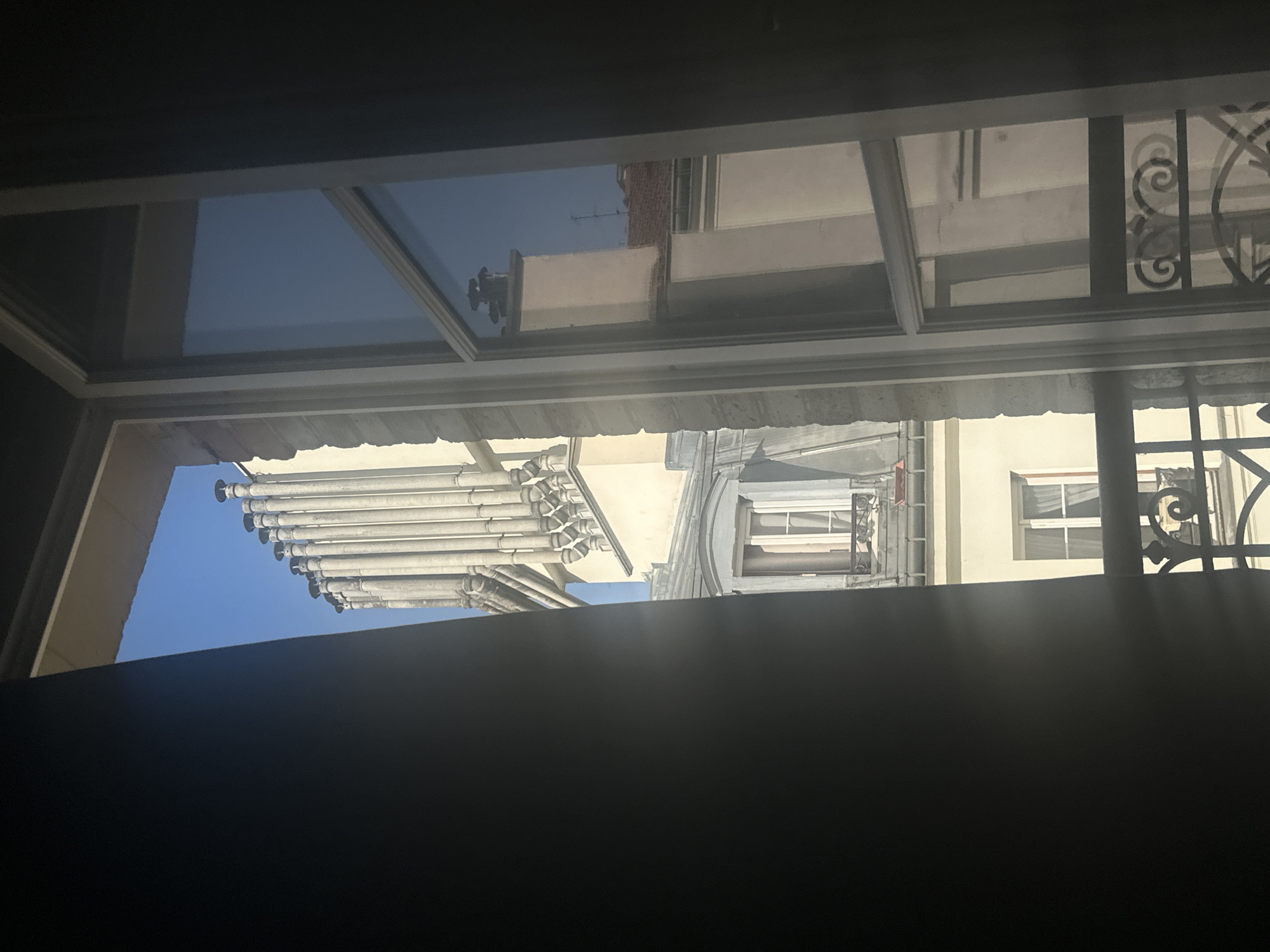 This is a photograph taken from inside an apartment in Paris, France, looking out through an open window. The view captures a classic Parisian rooftop scene, dominated by a row of tall, narrow chimneys on a light-colored building. The chimneys are arranged in a line and appear to be made of stone or concrete, with rounded caps. Below the chimneys, a building with a mansard roof is visible, featuring dormer windows with ornate iron railings. Another building with a cream-colored facade and white-framed windows can be seen further down.
The window frame itself is a prominent element, with the left side of the image showing a dark, silhouetted curtain or wall, creating a strong contrast with the bright outdoor scene. The right side of the image features the window pane, which reflects the blue sky and some of the buildings. The reflections are slightly distorted by the glass. Sunlight is streaming through the window, casting shadows and highlighting certain architectural details. The overall impression is one of a sunny day in Paris, with a view characteristic of its urban landscape. No people are visible in the image.
