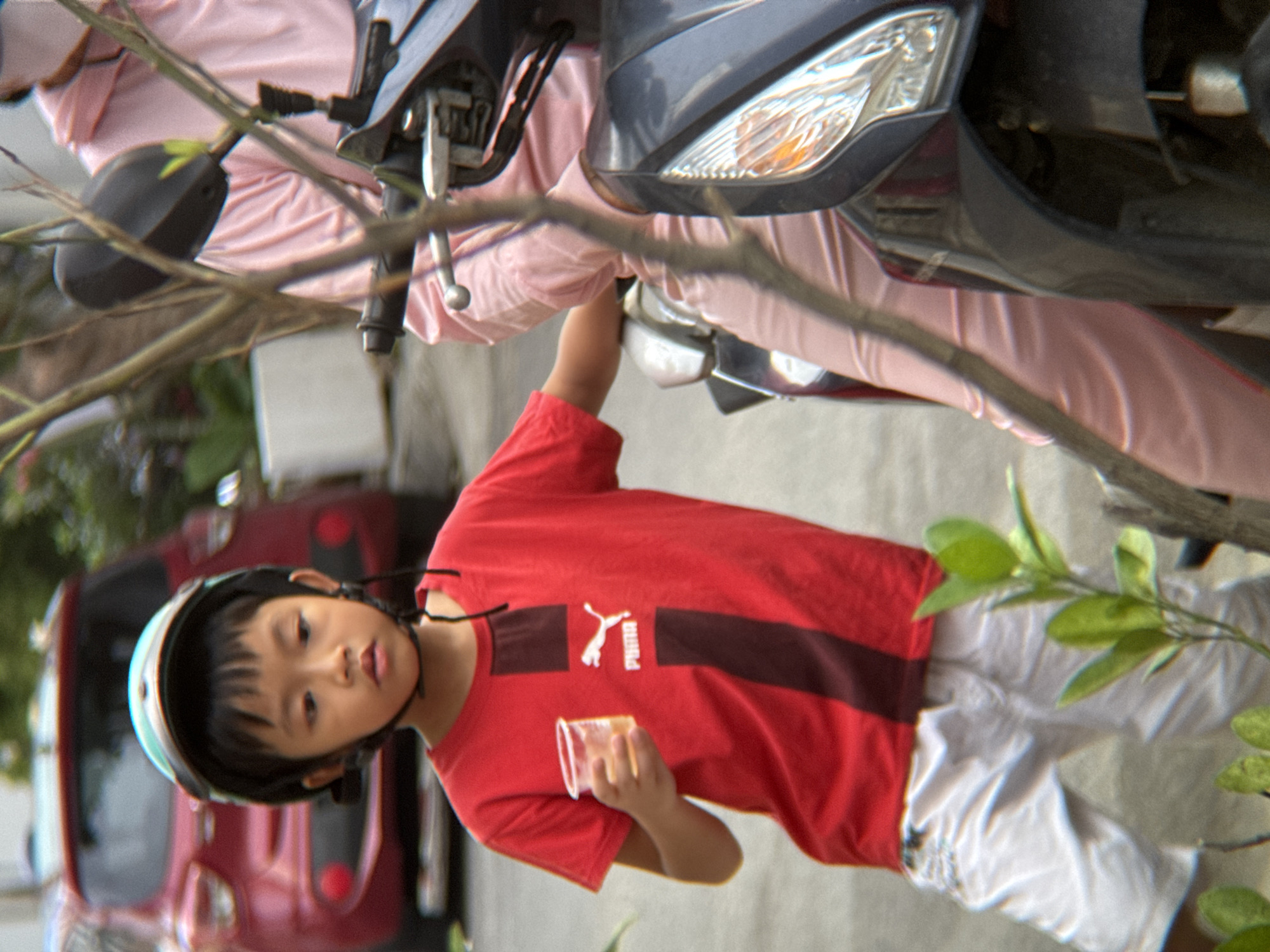 This media file captures a young boy, likely of Vietnamese descent given the location context of Nha Trang, Vietnam, as he is partially obscured by branches and foliage. He is wearing a red t-shirt with the Puma logo and a dark helmet. He is holding a clear plastic cup in his right hand. The boy is positioned near a motorbike, with parts of its dark gray body and a silver headlight visible. His red t-shirt has a horizontal stripe across the chest, featuring the white Puma leaping cat logo and the word "PUMA." He is also wearing white shorts. In the background, a red vehicle is partially visible. The lighting suggests it is daytime. The overall impression is that of a candid snapshot of a child outdoors, possibly on or near a street given the presence of vehicles and the slightly blurred background.