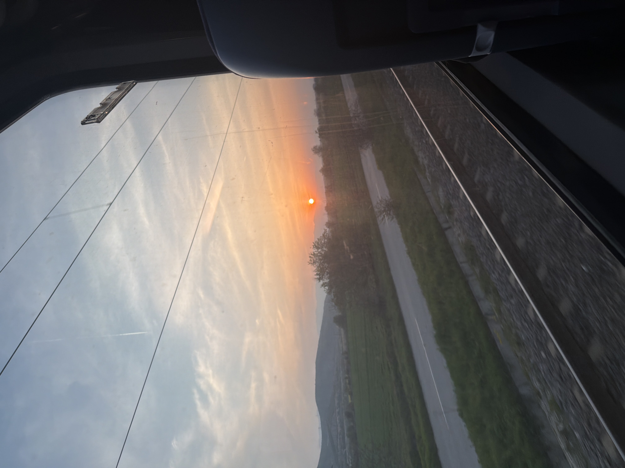 The image shows a landscape as viewed through the rear window of a car. The sun is setting, casting a warm orange glow on the sky, which fades into blue in the upper portion. A distant mountain range is visible. There is a road and green fields running alongside what appears to be a high stone wall or large wall of concrete blocks. There are defogger lines in the glass. At the top of the frame are parts of the car’s interior – sun visor, upper frame, and some object attached to the window. The location, Plovdiv, Bulgaria, cannot be confirmed solely from the image.