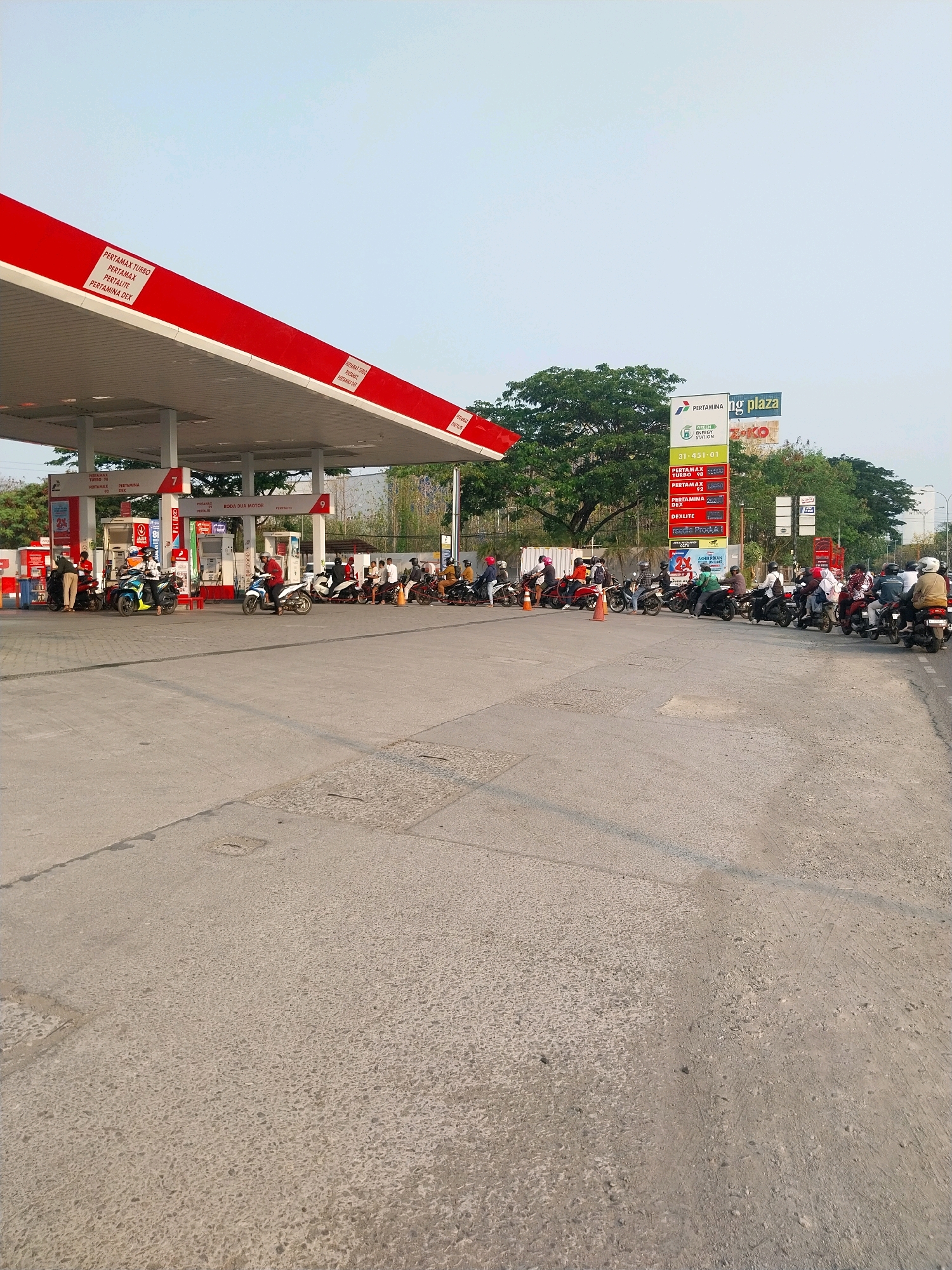 The image depicts a busy outdoor scene at a Pertamina gas station in Cirebon, Indonesia. The station features a red and white canopy over multiple fuel pumps. A long line of motorcycles, each with a rider, are queued up waiting to refuel. Some riders are seated on their motorcycles, while others stand nearby. Several attendants in uniform are visible near the pumps, some interacting with customers. A price board displaying different fuel types like "Pertamax Turbo," "Pertamax," and "Dexlite" is prominently displayed. In the background, trees and buildings, including a sign for "Ng Plaza," can be seen. The ground is paved concrete. The overall atmosphere suggests a typical day at a well-frequented gas station.