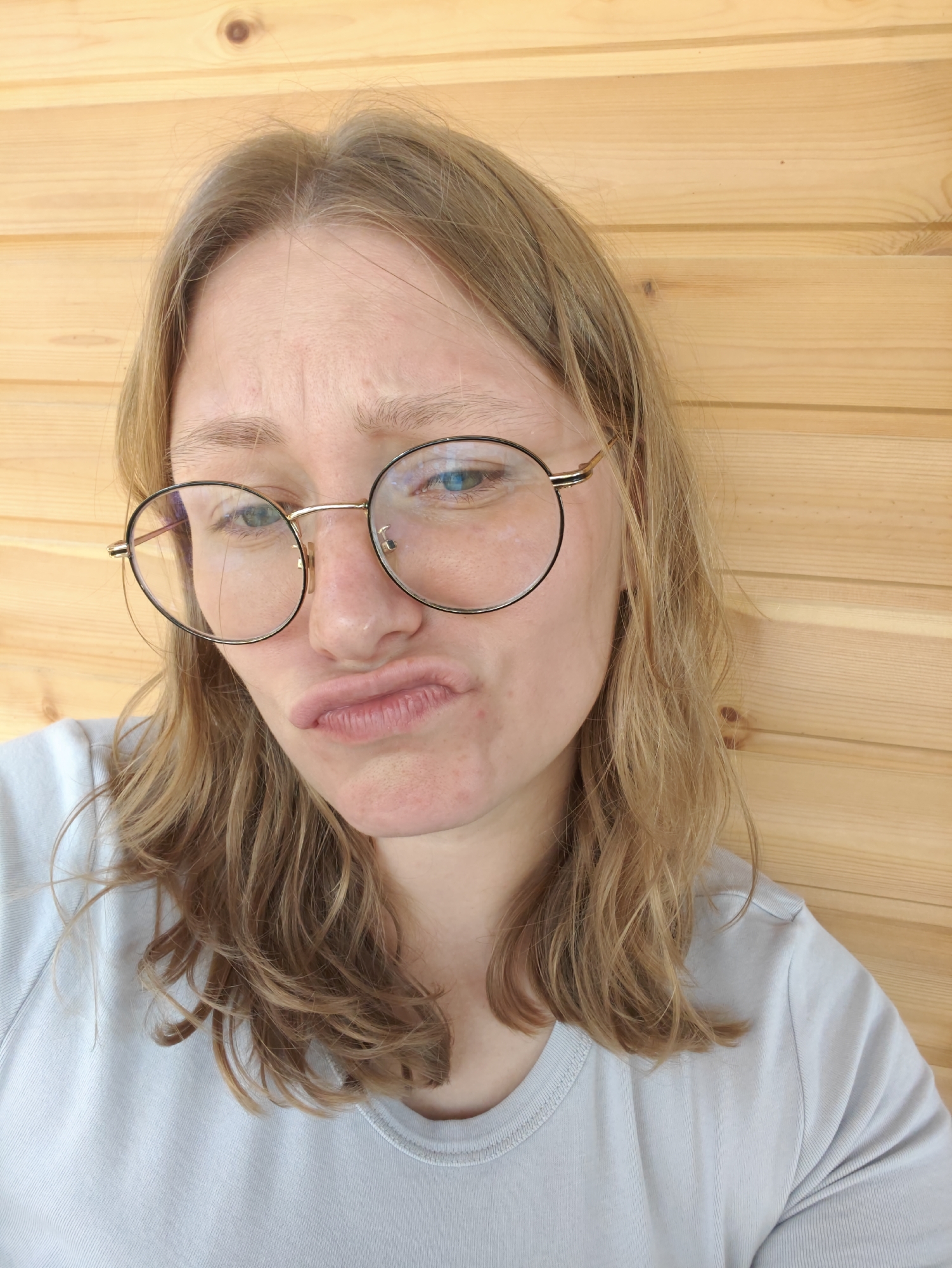 A young woman wearing round glasses and a light blue t-shirt stands in front of a wooden wall, her lips pursed in a comical expression. Her blonde hair is shoulder-length and slightly wavy, and her skin has a few blemishes. The lighting suggests an indoor environment, possibly a home or a studio. No specific location within Podolsk, Russia, can be identified from the image.