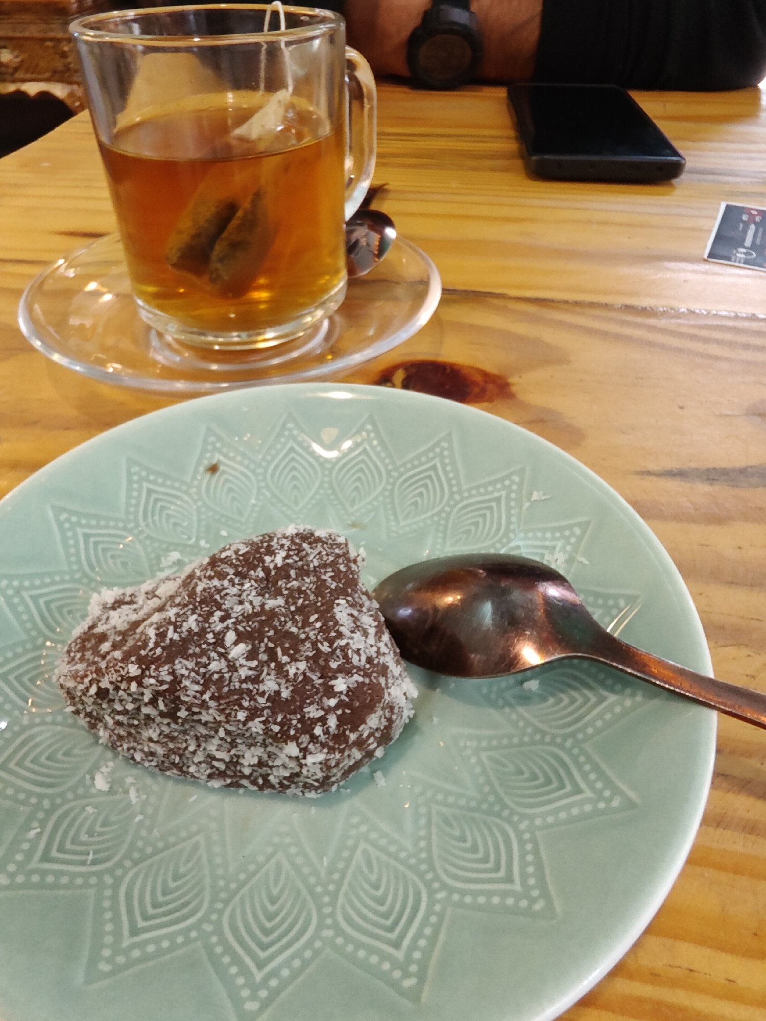 A close-up, eye-level shot captures a cozy cafe scene in Vashisht, India. In the foreground, a light blue, circular plate with an intricate white mandala design holds a single, heart-shaped chocolate cake coated in shredded coconut. A copper-colored spoon rests to the right of the cake. Behind the plate, a clear glass mug filled with amber-colored tea and two tea bags sits on a clear glass saucer. The mug and saucer are placed on a light brown wooden table, which is also occupied by a black smartphone, a small white card with black text, and a person's wrist wearing a dark watch. The overall ambiance is relaxed and inviting, typical of a cafe setting.