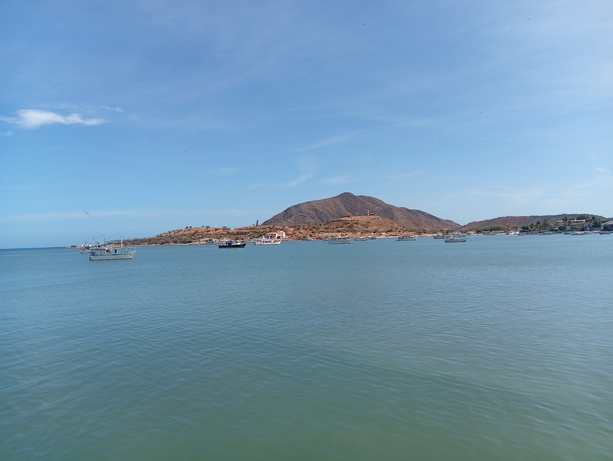The image depicts the harbor of Juan Griego, Venezuela, on a clear day. The scene includes a body of water, likely the Caribbean Sea, with gentle ripples across the surface. Several boats are anchored in the harbor at varying distances. In the background, a prominent, brown, dry mountain rises from the landscape. Some structures and vegetation can be seen along the mountain's base and extending towards the water. The sky is blue with a few scattered clouds and numerous birds flying around.