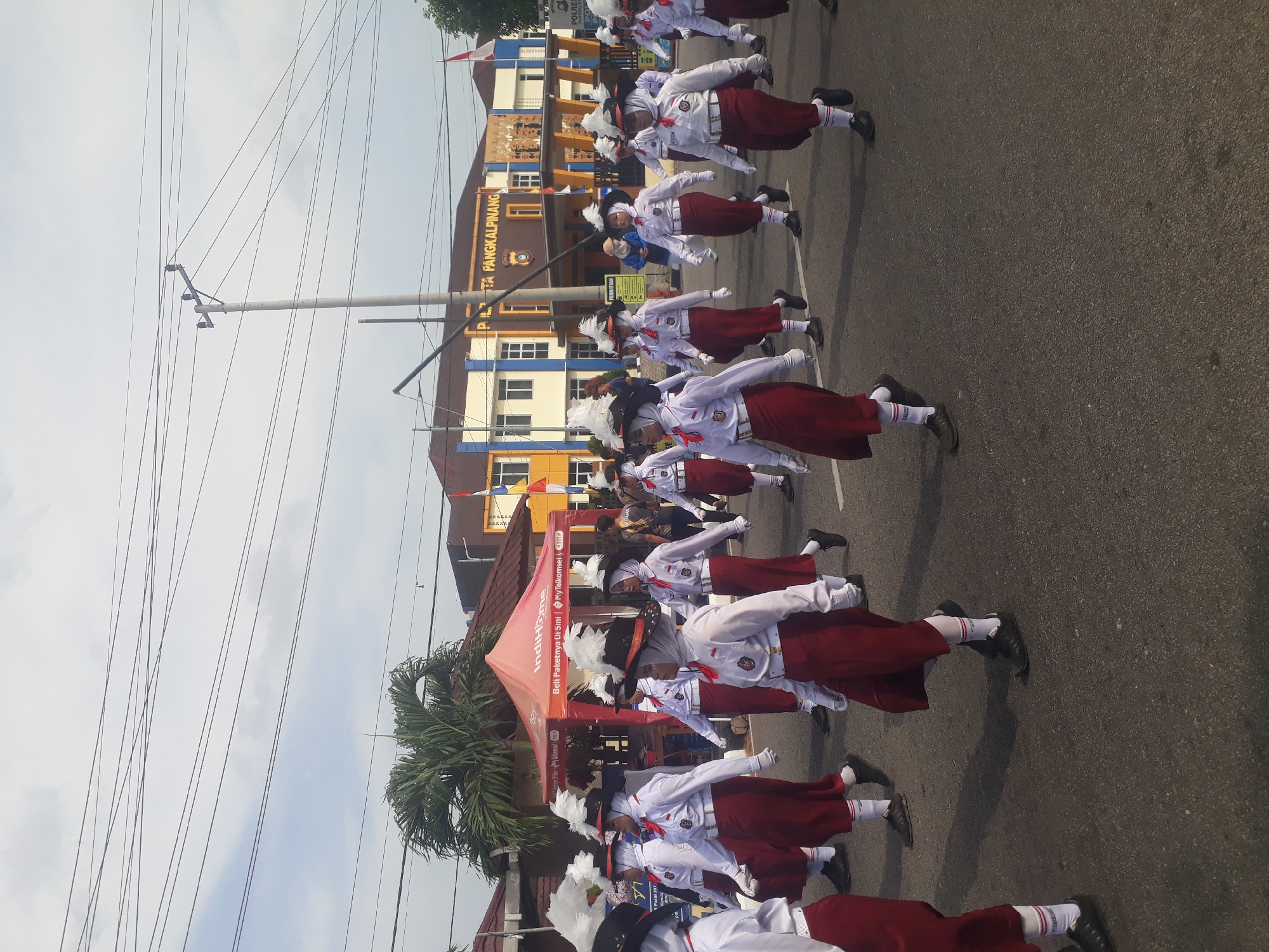 A group of young people in uniform are marching in a parade. They are wearing white shirts with red collars and cuffs, white hijabs, and red skirts or shorts. They also have black hats with white feathers and white gloves. They are marching on a paved road in front of a building that has "POLRES PANGKALPINANG" written on it, indicating the location is in Pangkalpinang, Indonesia. The sky is cloudy. There are power lines and a palm tree visible in the background. A red tent with "IndiHome" branding is set up on the side of the road.