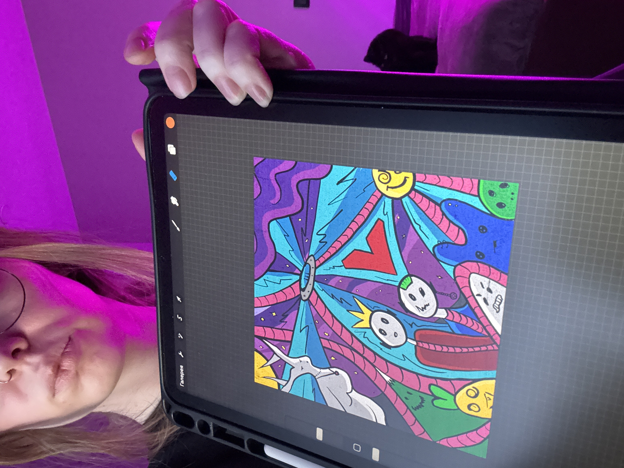 An indoor, close-up photograph captures a person holding a tablet, showcasing a vibrant digital artwork. The person, likely female, is partially visible on the left side, with light-colored hair, glasses, and a nose piercing. Her face is illuminated by a strong magenta/purple light that dominates the background, suggesting mood lighting within a room in Krasnoyarsk, Russia. In the upper background, a dark, indistinct shape is visible, possibly a pet or object.
The main subject is a black tablet, held upright, displaying a colorful, intricate digital illustration. The artwork features a psychedelic or doodle-art style with a rich palette of purples, blues, pinks, reds, greens, and yellows. Abstract shapes, swirling lines, and cartoon-like elements fill the canvas, including a smiling yellow sun, skull-like faces, a green alien head, and other fantastical figures. The interface of what appears to be an art application (possibly Procreate) is visible on the left side of the screen, showing tools and the Russian word "Галерея" (Gallery), indicating the person is likely creating, viewing, or sharing their digital art. The person's expression is neutral as they hold the device.